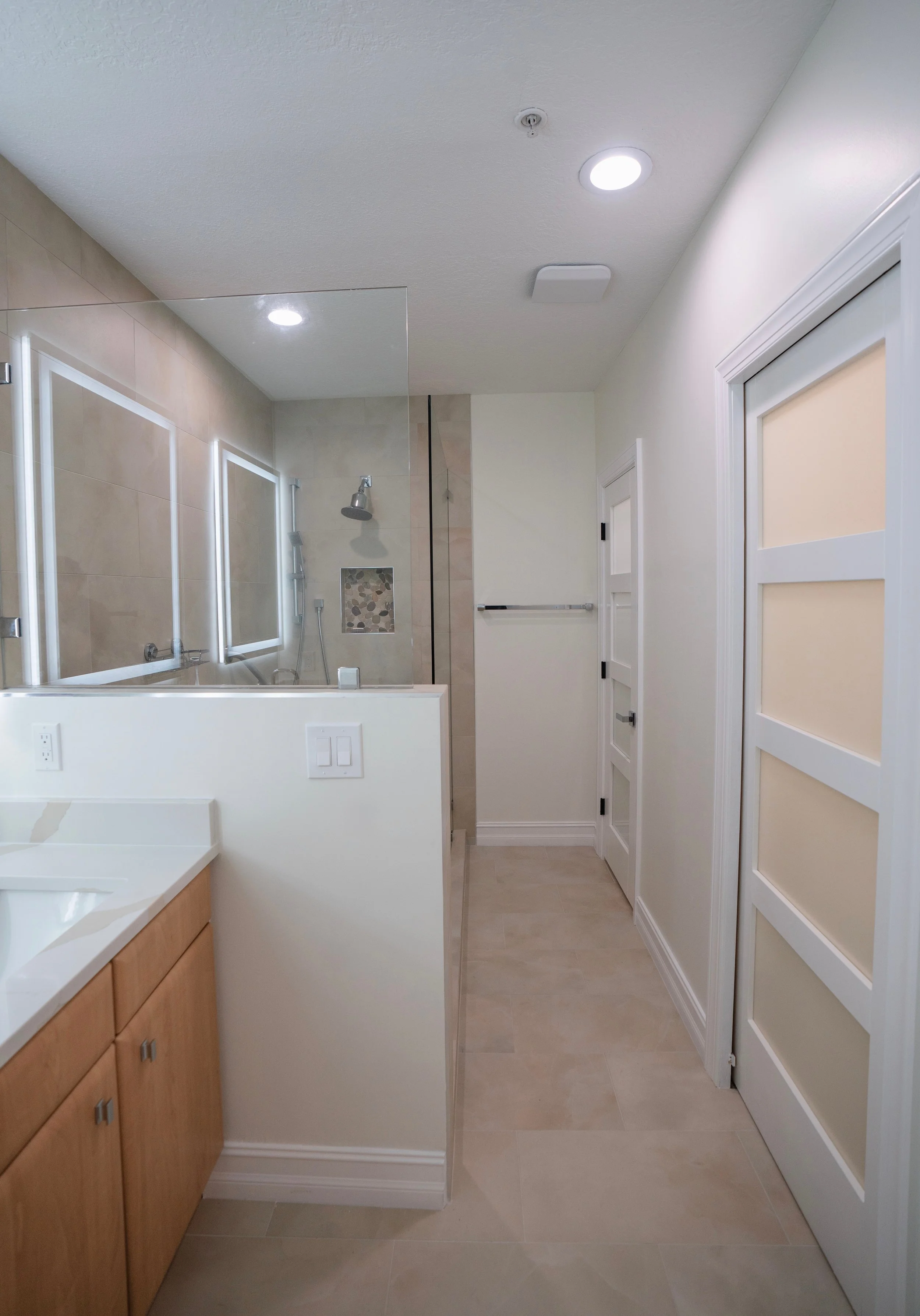 Modern bathroom with beige tiles, white walls, a wooden vanity, glass shower enclosure with a window, and ceiling lights.