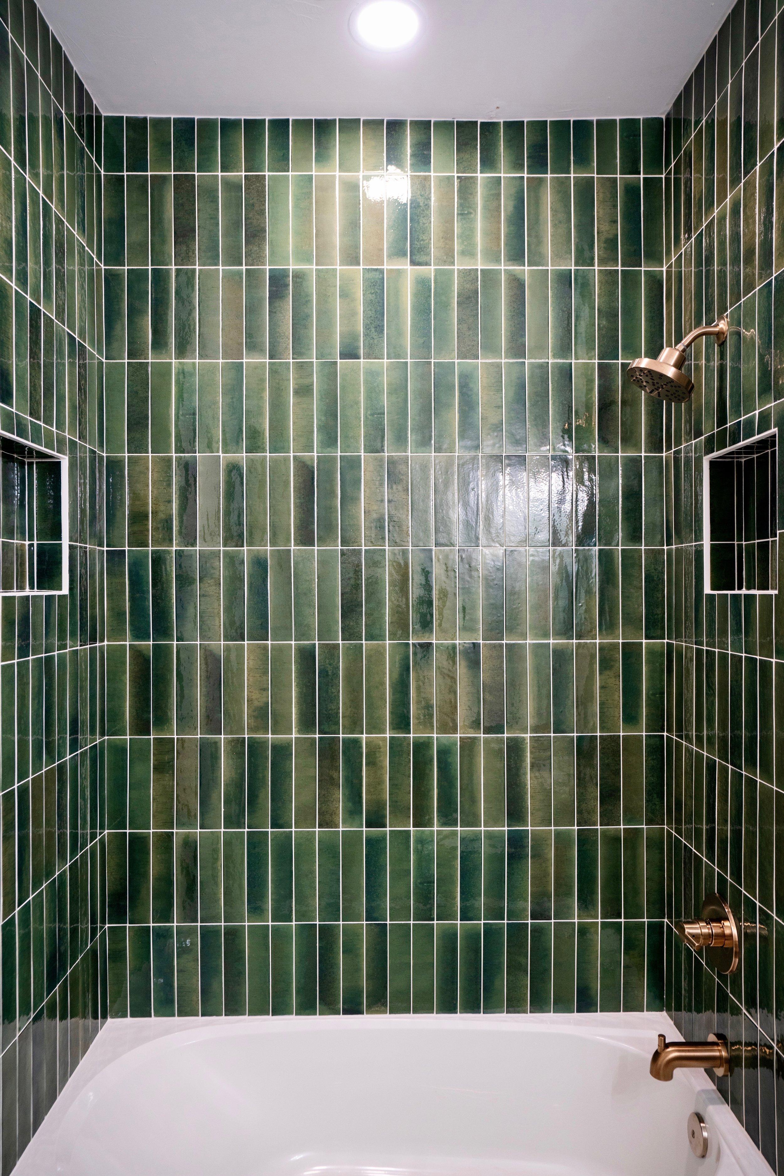 Green tiled shower with a bronze showerhead, faucet, and built-in wall niche, with a white bathtub at the bottom and a ceiling-mounted light.