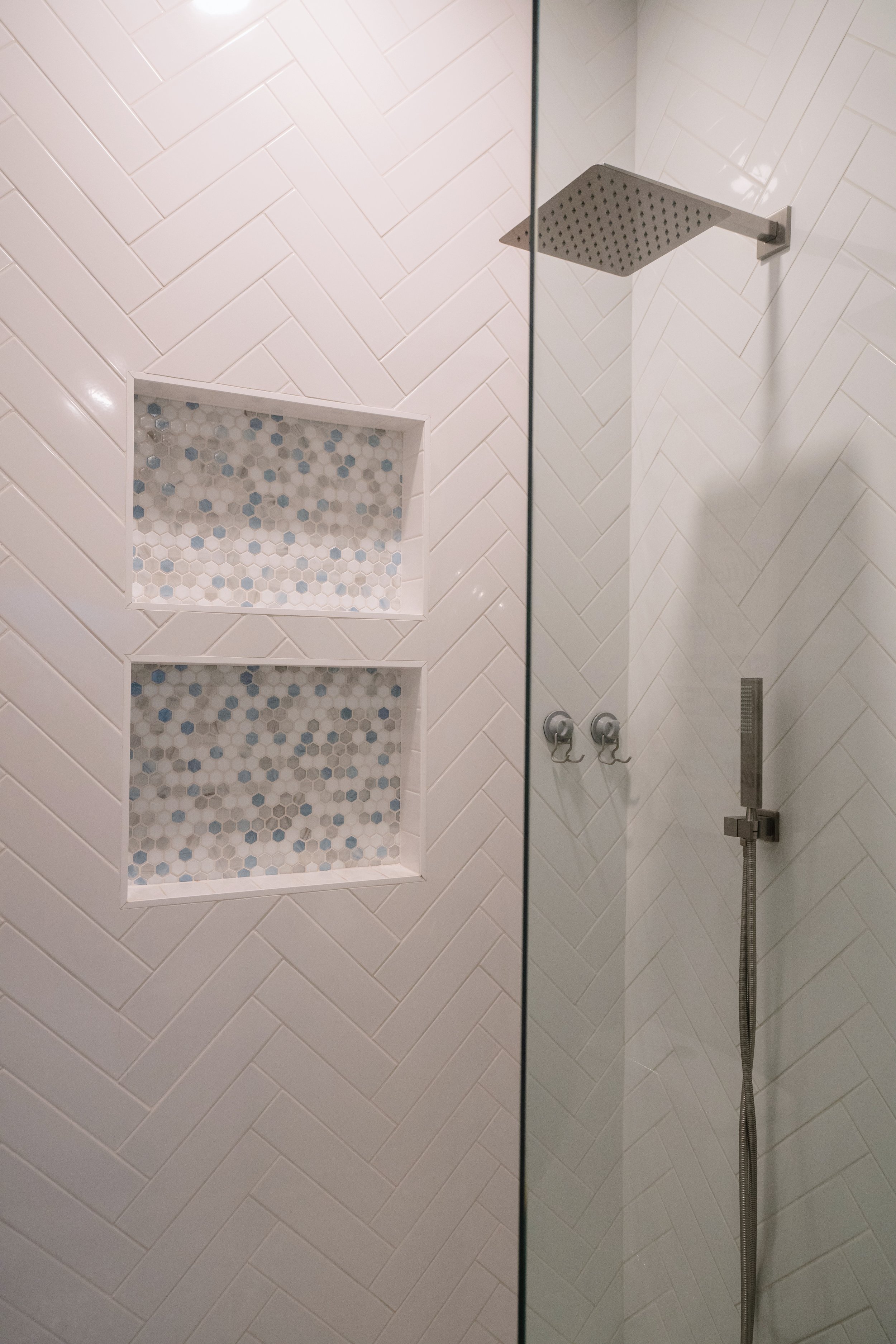 Detail of a modern glass shower wall, showing a rain showerhead, handheld shower, wall-mounted controls, and built-in niches with hexagonal tile pattern.