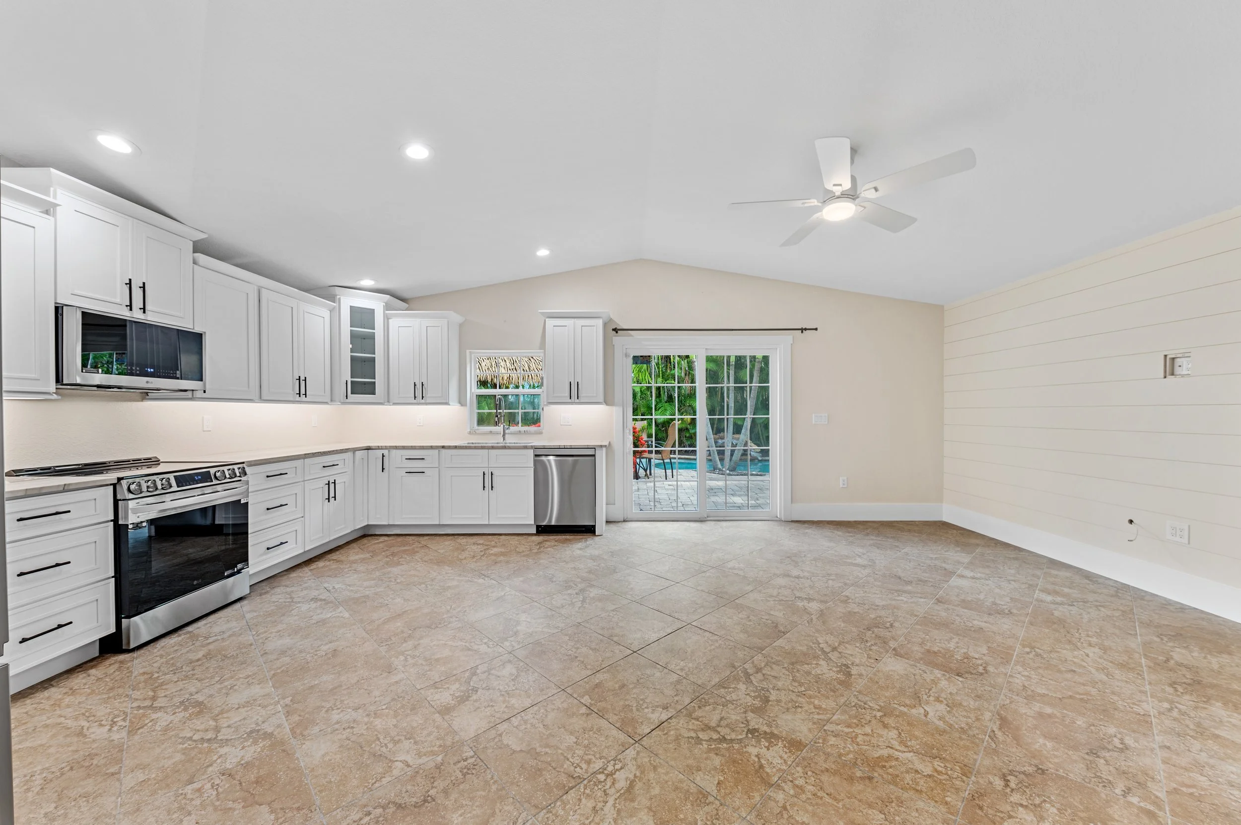 Empty kitchen and living room area with beige tiled floor, white cabinets, stainless steel appliances, sliding glass door leading to backyard with trees and patio furniture, ceiling fan, recessed lighting.