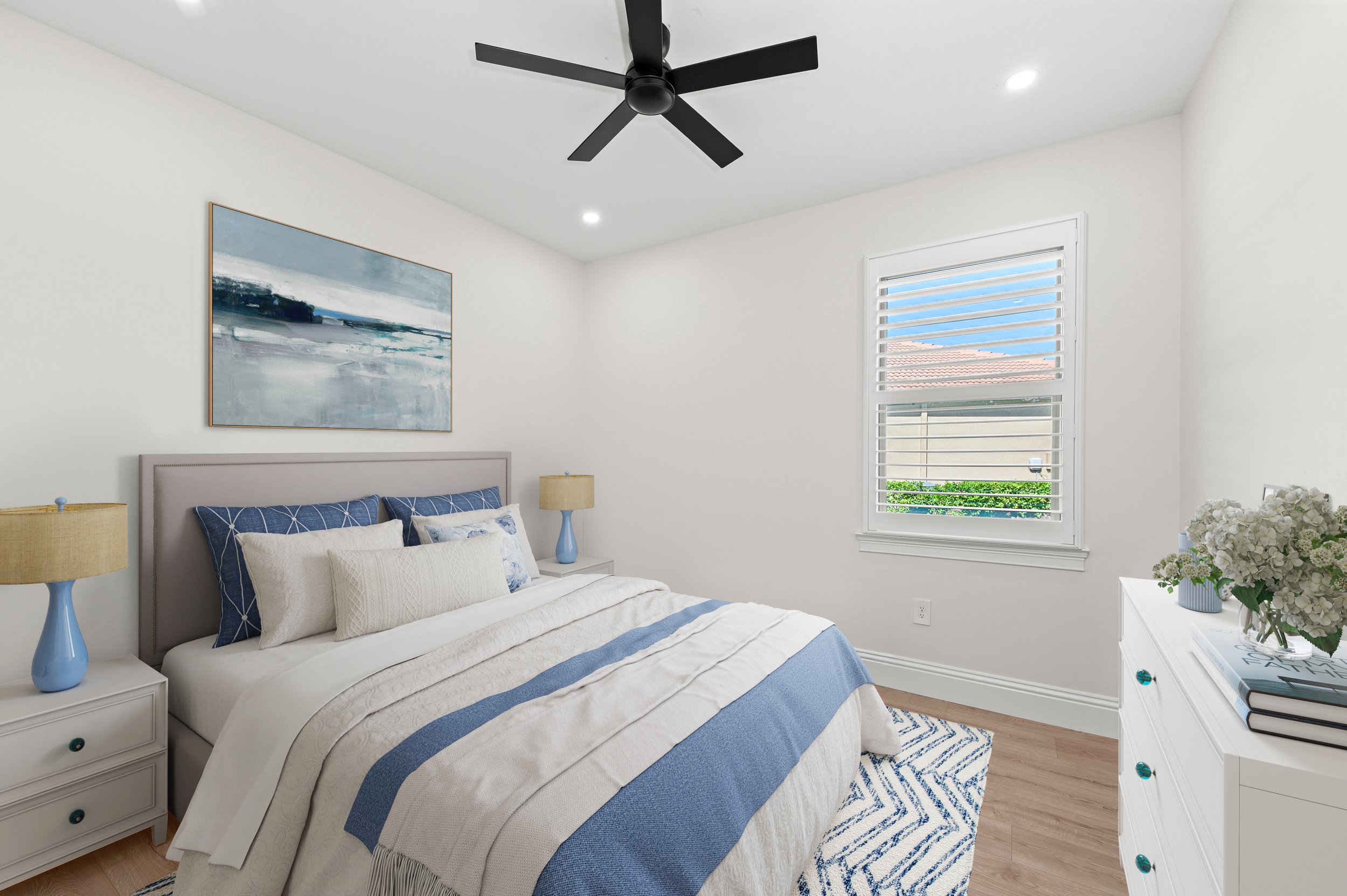 Bedroom with white walls, a large bed with blue and white bedding, two nightstands with blue lamps, a window with white blinds, and a white dresser with a vase of white flowers.