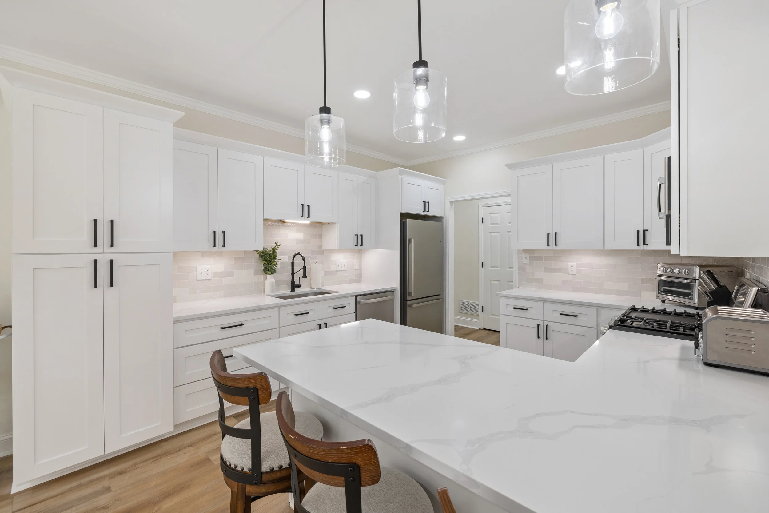 Modern white kitchen with marble island, black cabinet handles, pendant lights, stainless steel appliances, and wooden chairs.