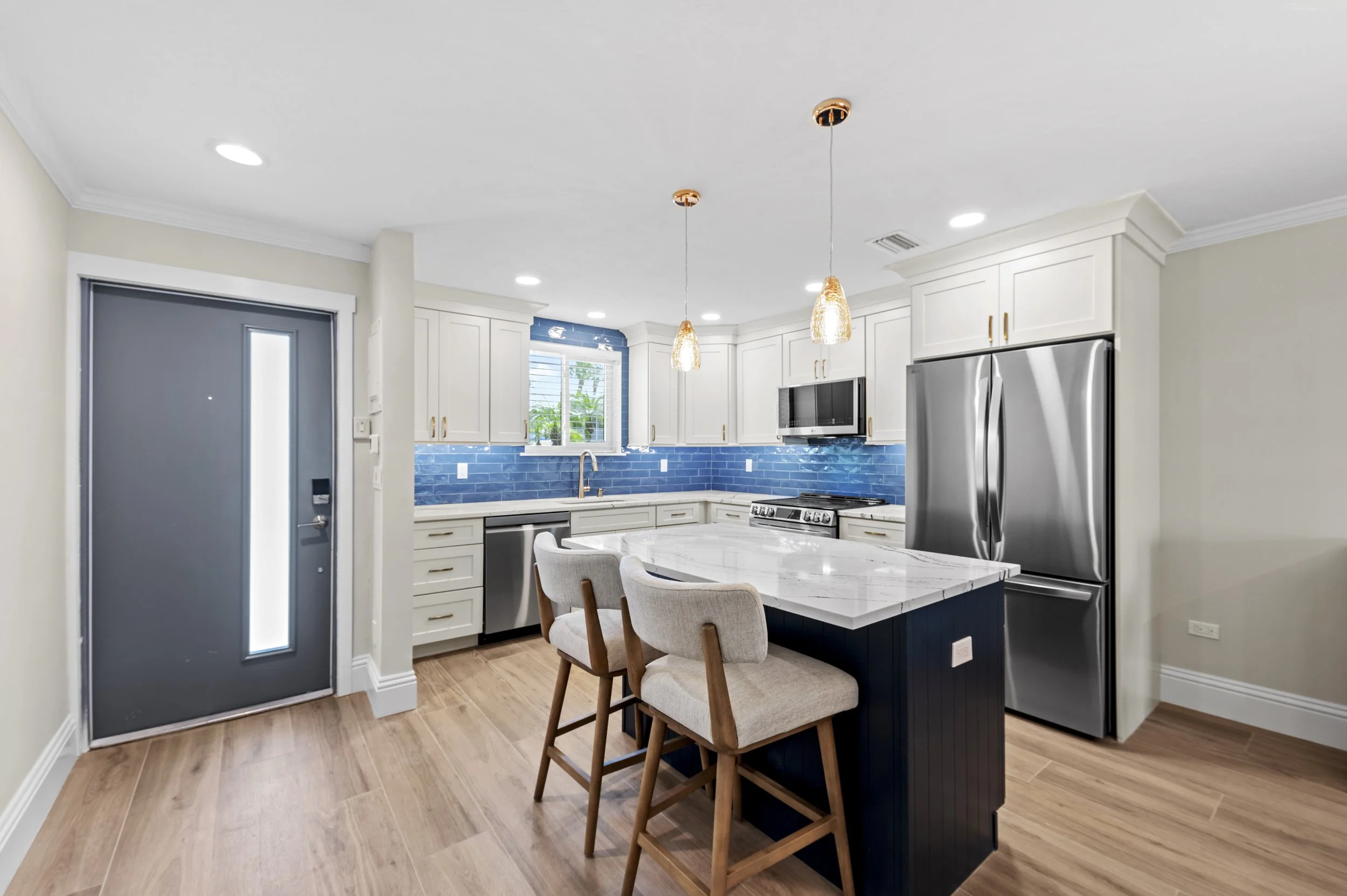 Modern kitchen with white cabinets, blue backsplash, stainless steel appliances, a marble island with seating, pendant lighting, and hardwood floors.