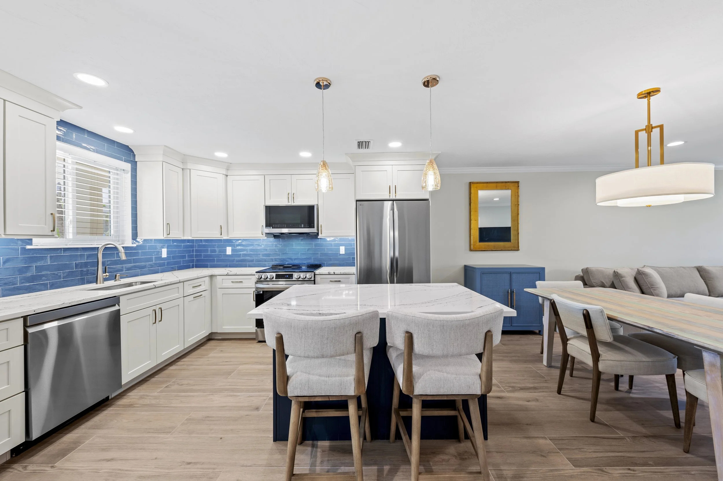 Open-concept kitchen with white cabinets, blue subway tile backsplash, stainless steel appliances, and a central white marble island with three cushioned chairs. Adjacent dining area has a wooden table with chairs, a beige sofa, a blue cabinet, and a