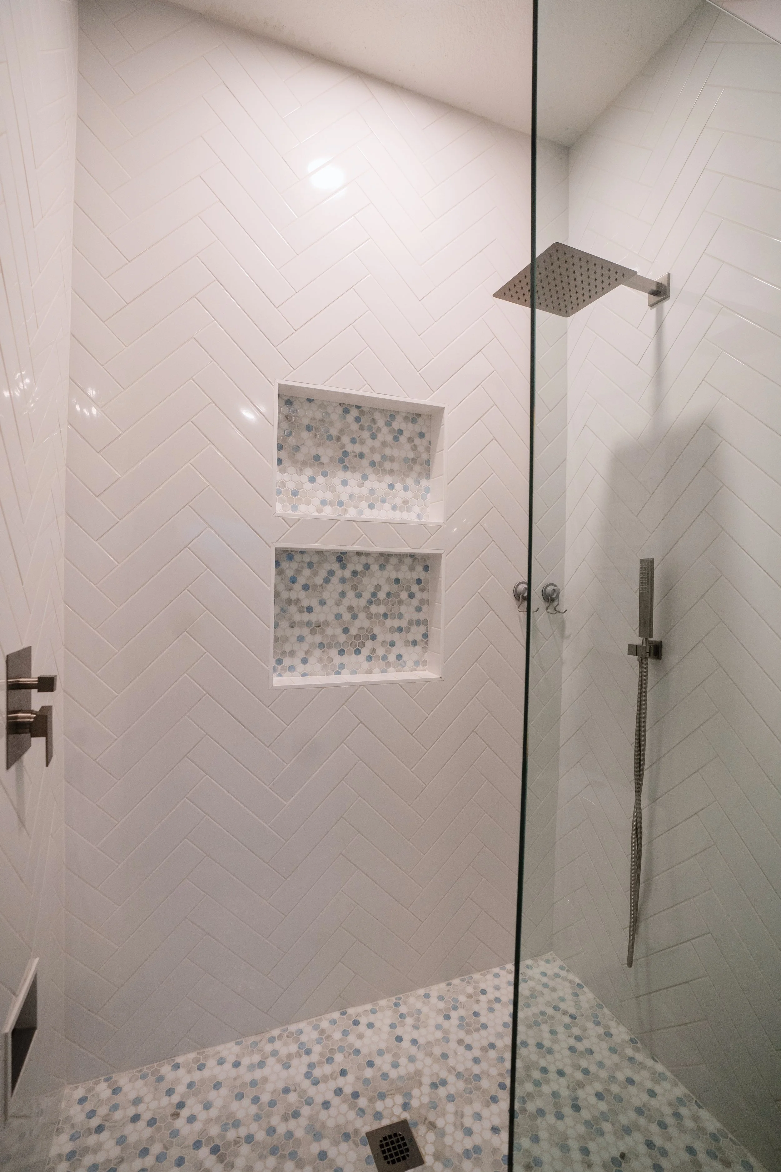 Modern walk-in shower with white herringbone tile walls, two built-in niche shelves with mosaic tiles, and a rain showerhead.