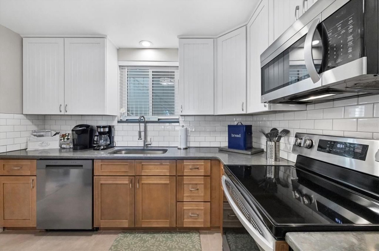 Kitchen with white and wooden cabinets, stainless steel appliances including a microwave, oven, dishwasher, and coffee maker, white subway tile backsplash, window above the sink, and various kitchen items on the counters.