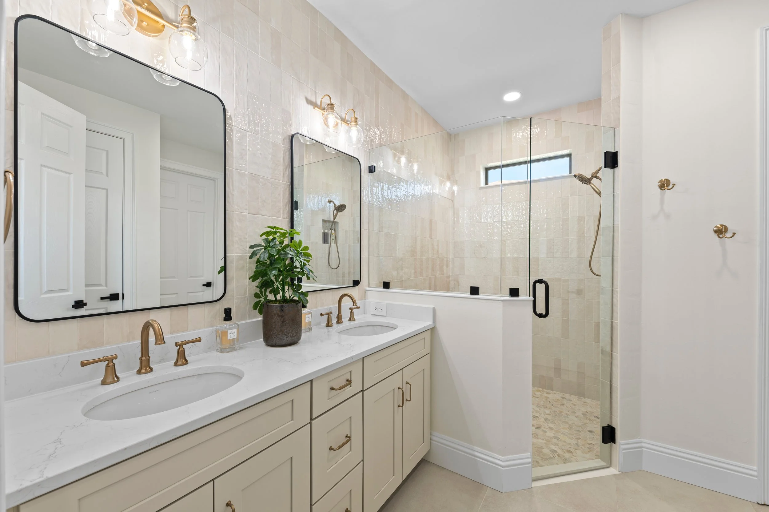 Modern bathroom with dual sinks, white countertop, gold fixtures, large mirrors, a green plant, wall-mounted lights, and a walk-in shower with glass doors and beige tiles.