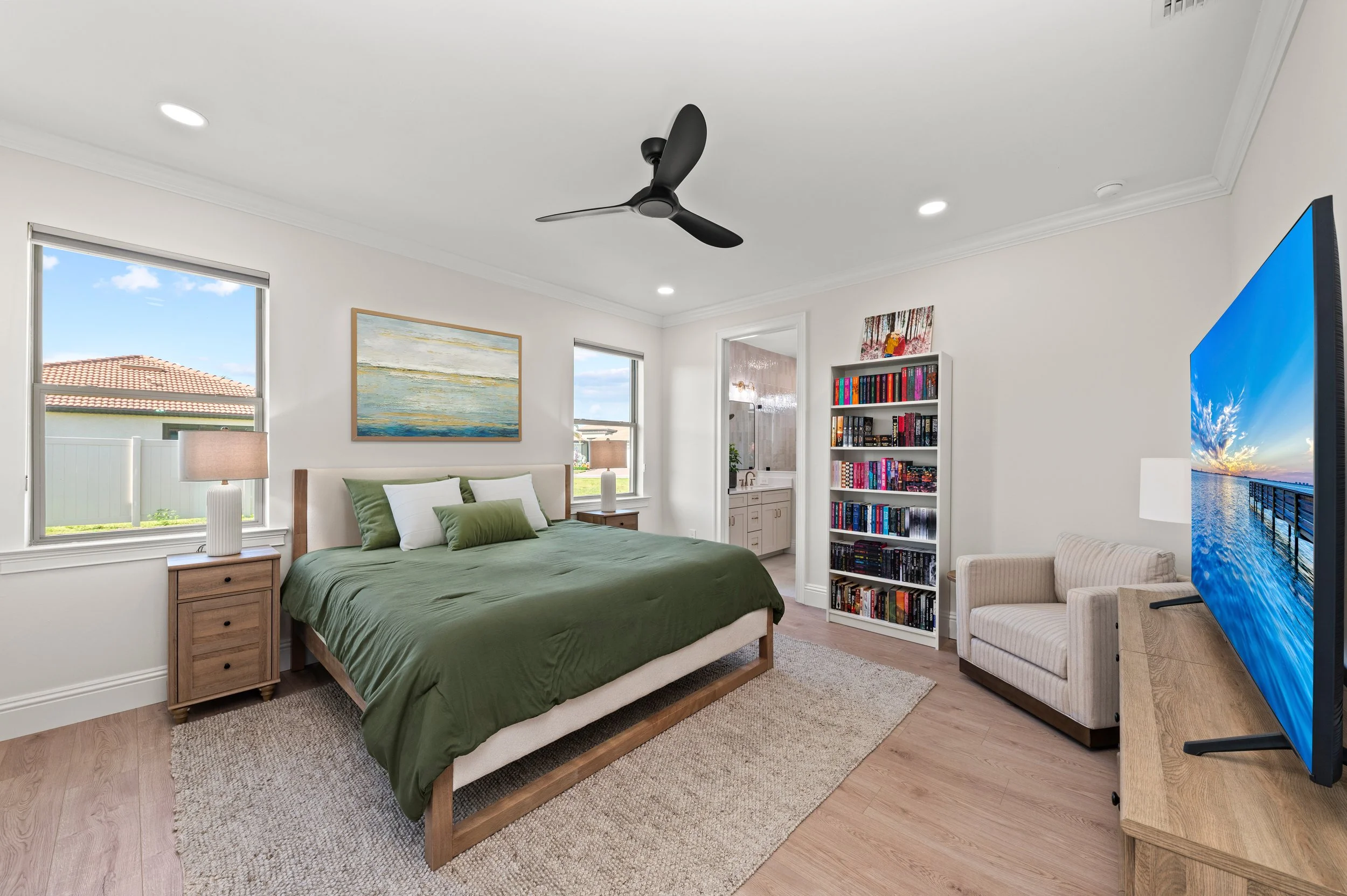Bedroom with green bedding, wooden nightstand, table lamp, two windows, a bed, bookshelf filled with books, a beige armchair, a large flat-screen TV, and an en suite bathroom.