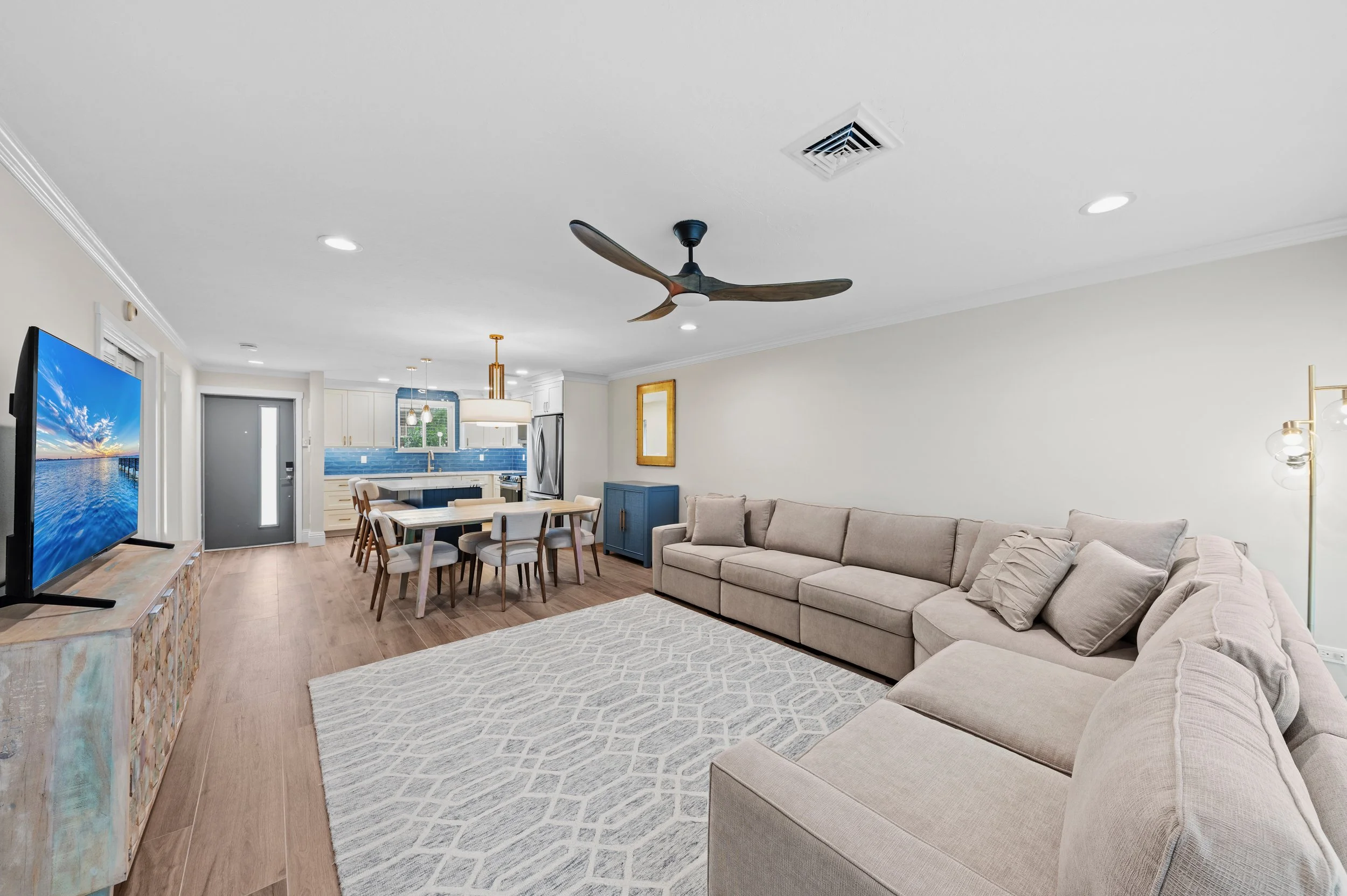 Open living room with a beige sectional sofa, a white geometric rug, a large flat-screen TV on a wooden media stand, and a ceiling fan. The background shows a dining area with a wooden table and chairs, and a kitchen with white cabinets, blue backspl