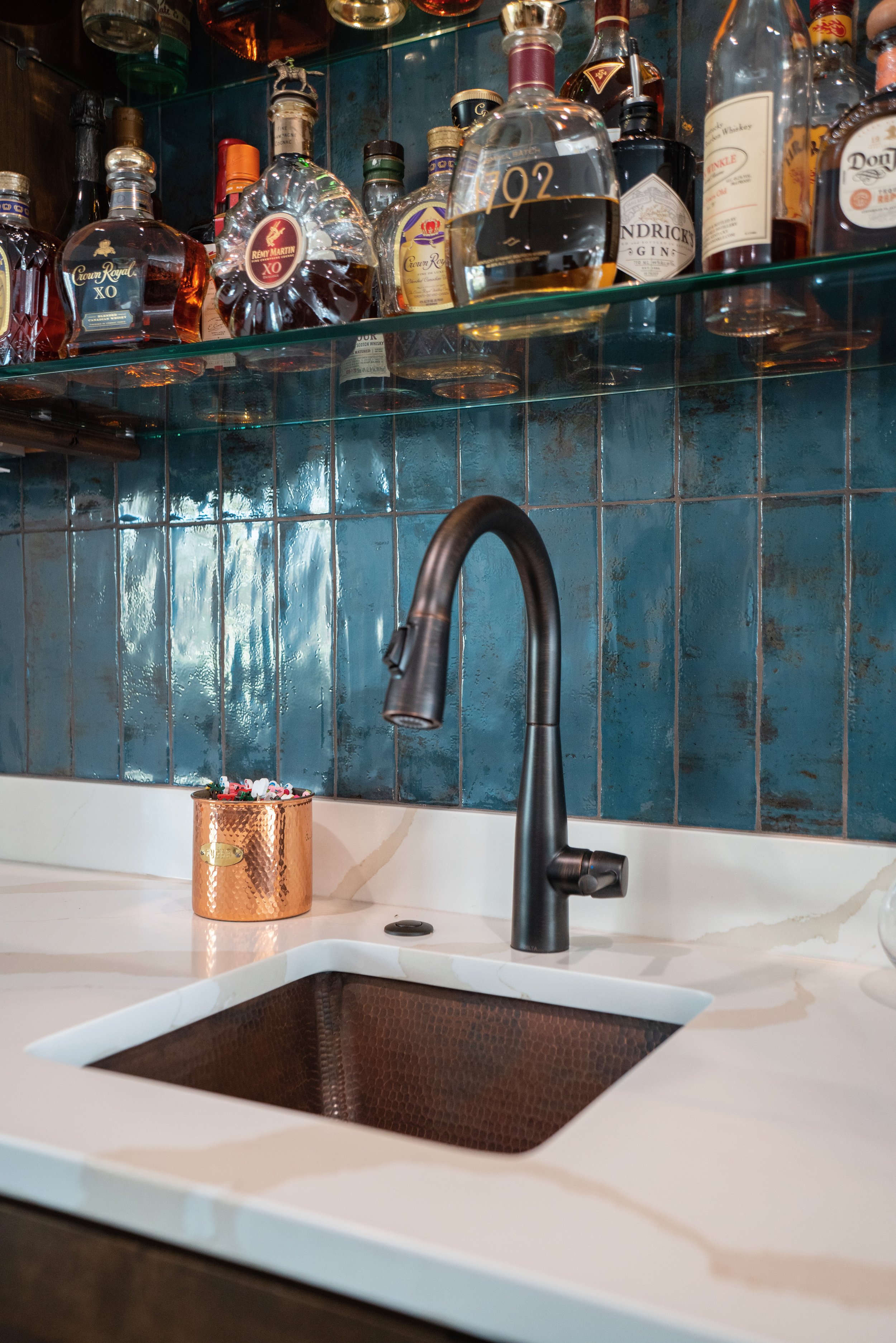 A kitchen sink with a black faucet, a white marble countertop, blue tiled backsplash, a copper-colored cup, and a shelf with various liquor bottles including Crown Royal, Rémy Martin XO, Hendrick's Gin, and others.