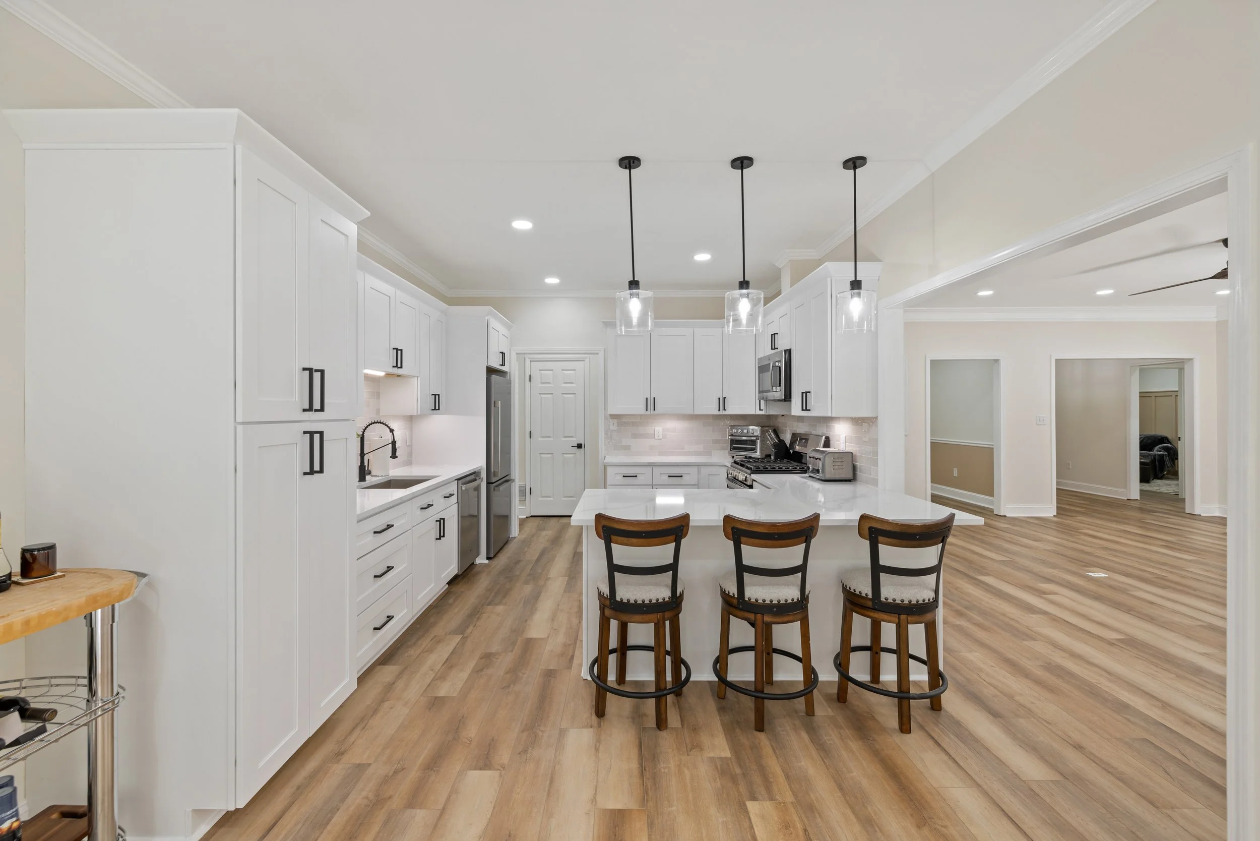Modern kitchen with white cabinets, a central island with a white countertop, three bar stools, stainless steel appliances, and pendant lights hanging above the island.