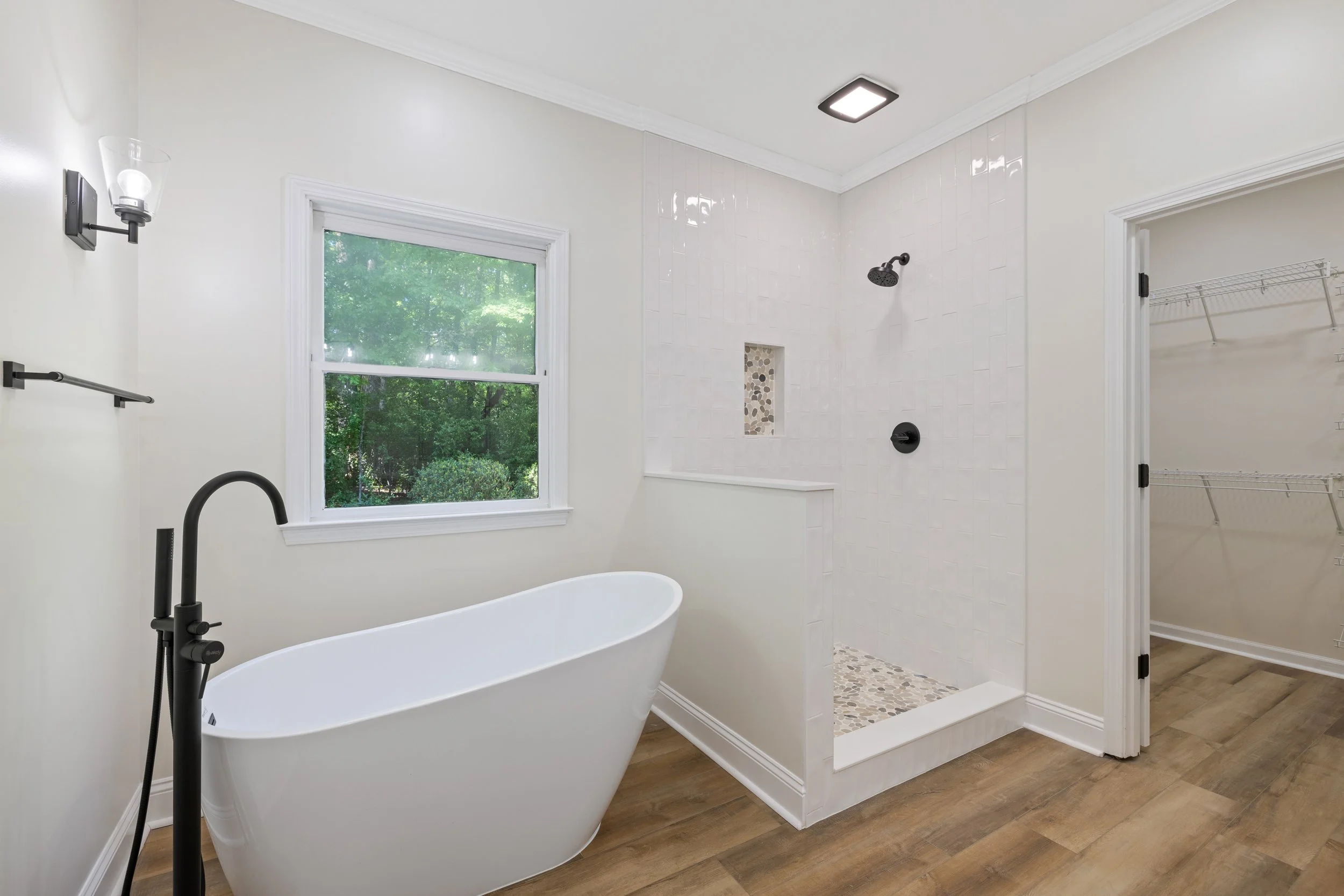 Modern bathroom with a white freestanding bathtub, black fixtures, large window overlooking greenery, and a walk-in shower with white tile and pebble floor, adjacent closet with wire shelving.