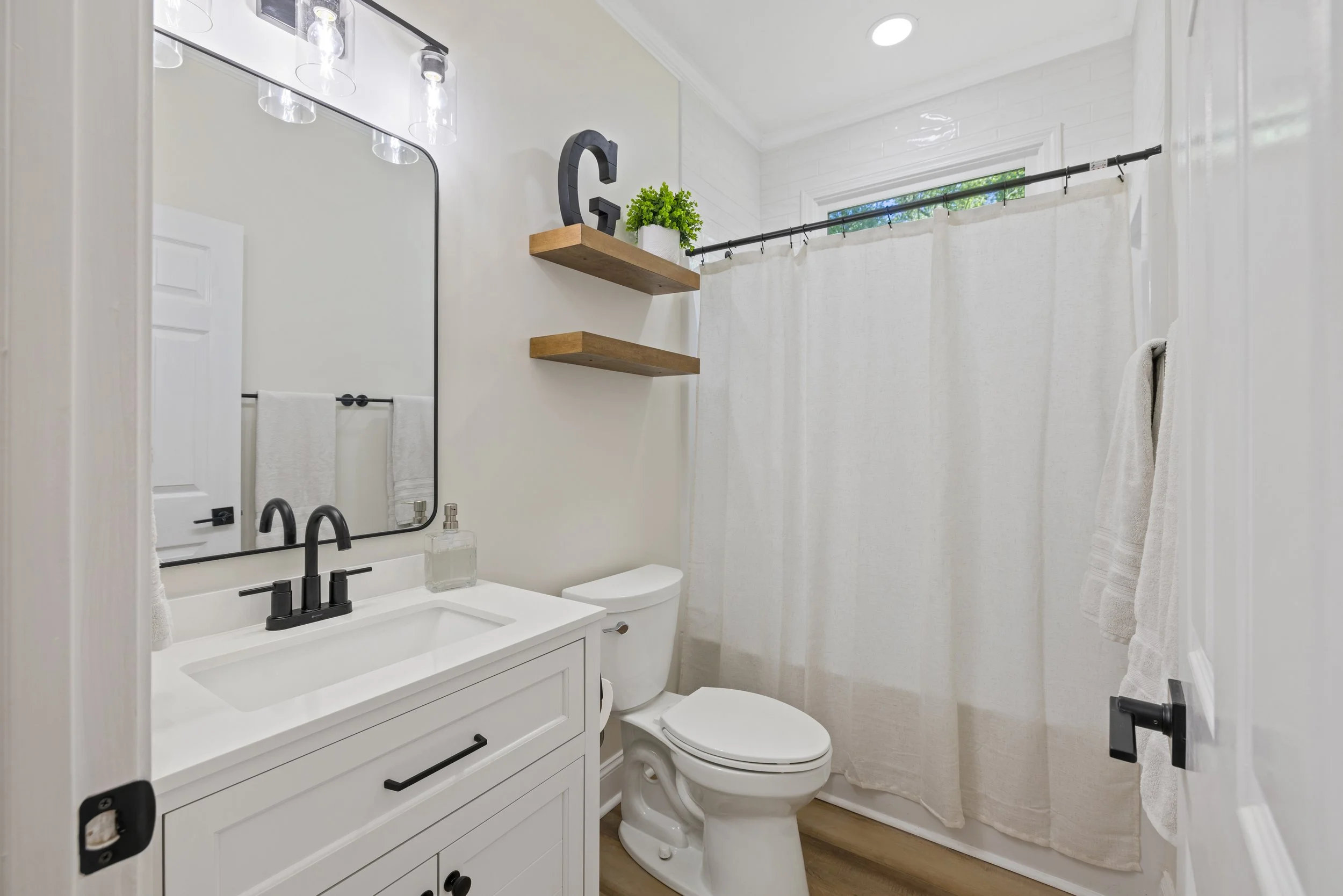 A small bathroom with a white vanity, black faucet, and large mirror, a toilet, two wooden shelves with a green plant and a decorative letter G, and a shower with a white curtain and a window with greenery outside.