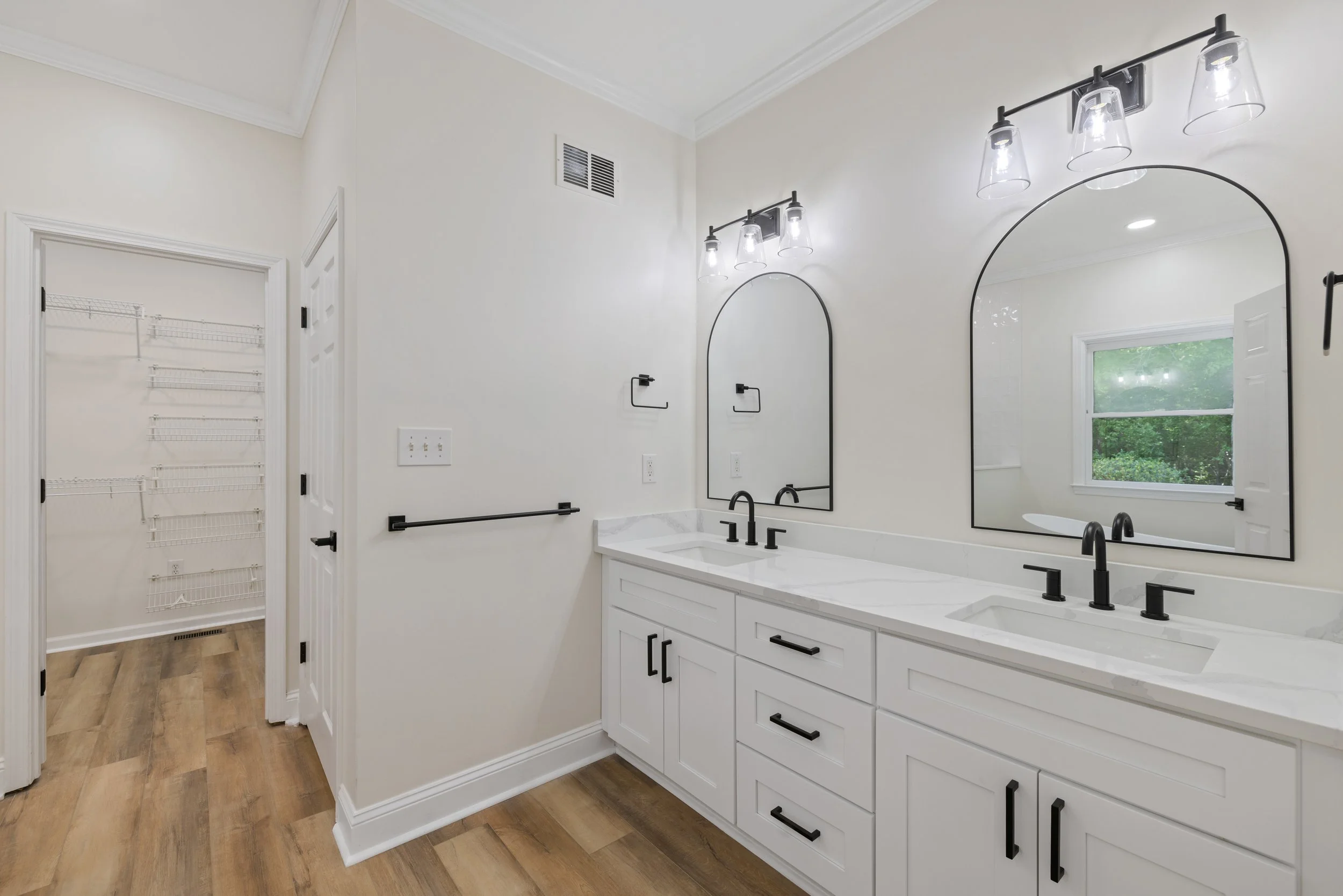 Modern bathroom with white vanity, black fixtures, large arched mirrors, and wood flooring. Includes a window overlooking greenery and a walk-in closet with wire shelving.