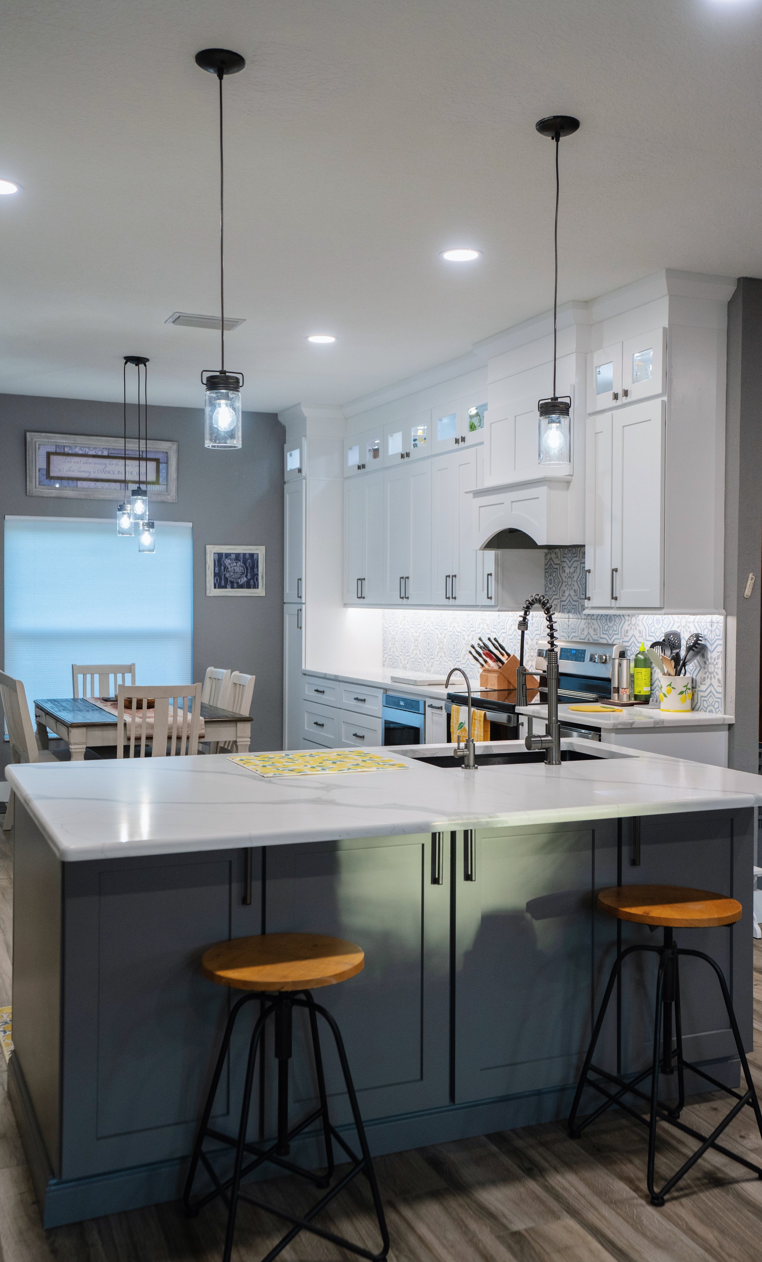 Modern kitchen with white cabinets, white marble countertop, and black fixtures. Central island with gray base and wooden bar stools. Dining area with a light-colored table and white chairs in background. Pendant lights above island, recessed lightin