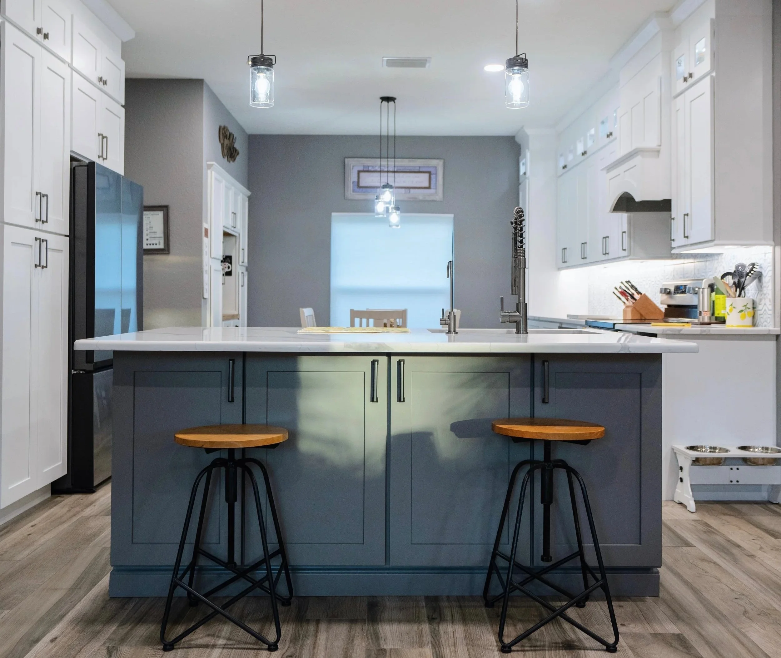 Modern kitchen with a gray island, white cabinets, and wooden bar stools, illuminated by pendant lights.
