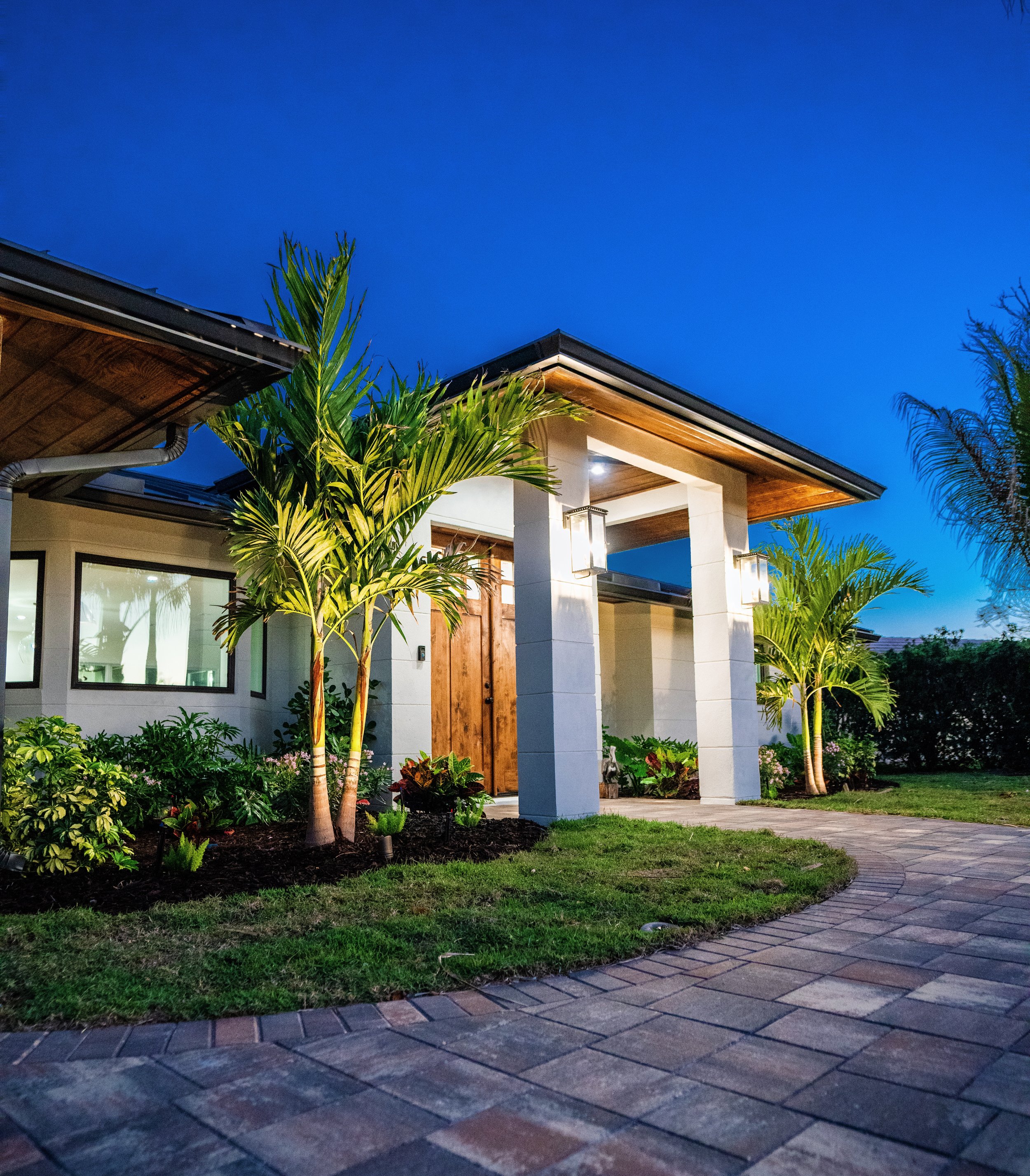 Modern house entrance with stone pillars, wooden door, tropical plants, and front pathway at dusk.