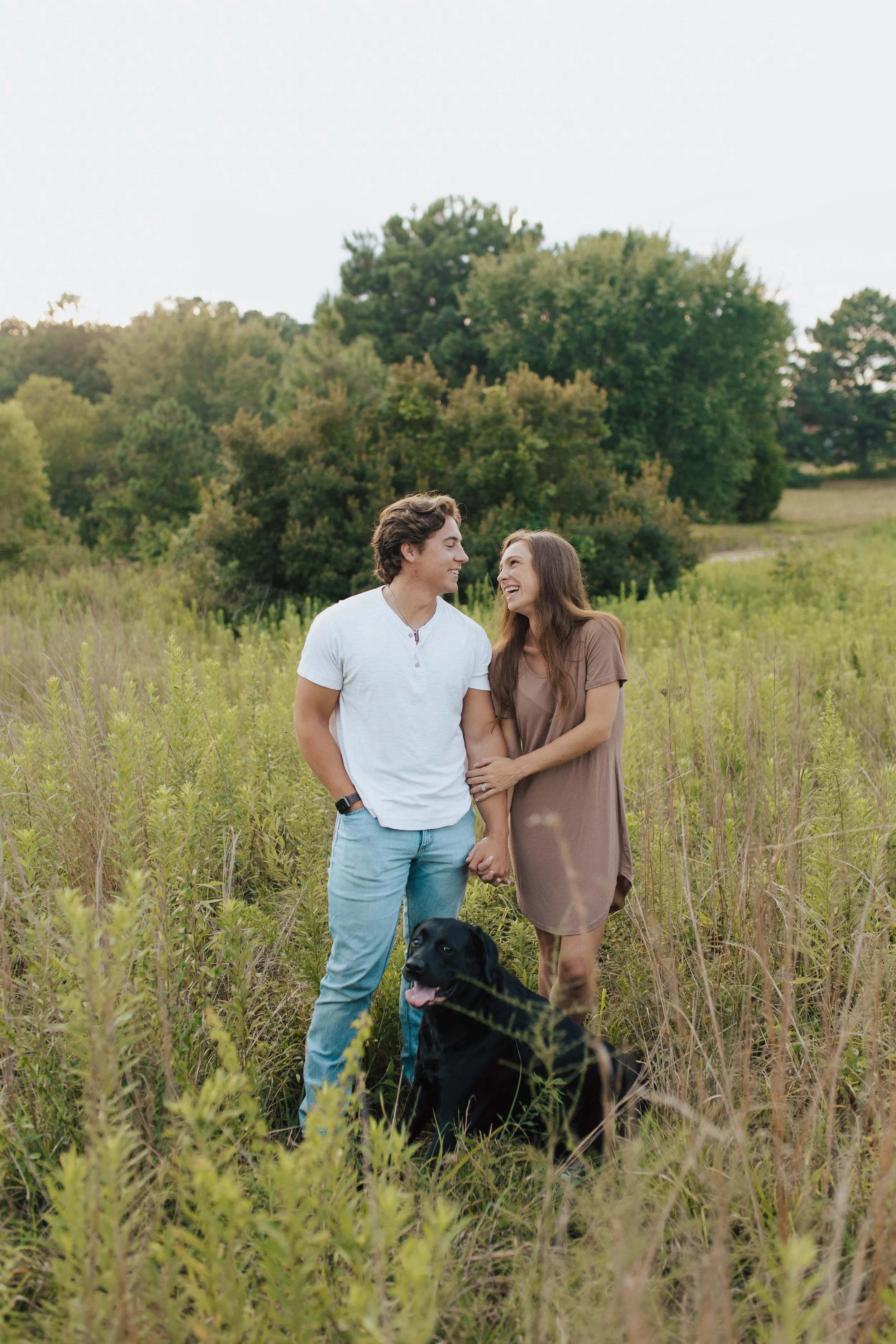 A young couple holding hands and smiling at each other in a grassy field with a black dog sitting nearby, surrounded by trees.