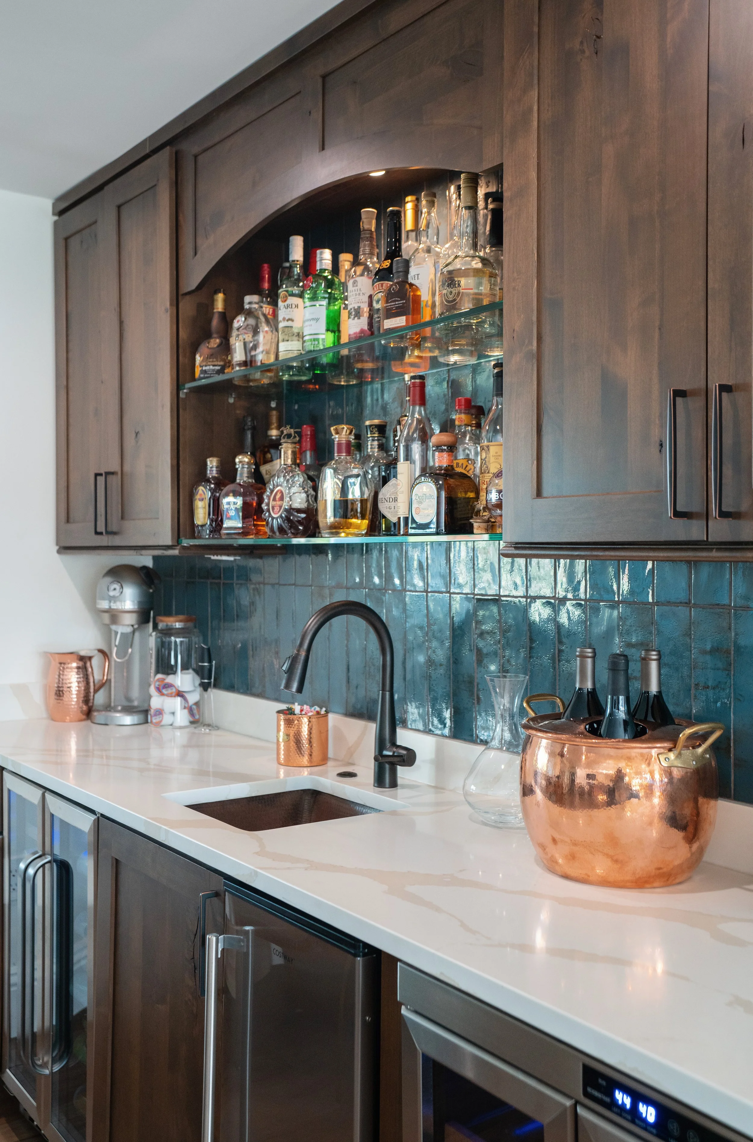 Home bar area with wooden cabinets, shelves of assorted liquor bottles, copper buckets, a sink, and a marble countertop.