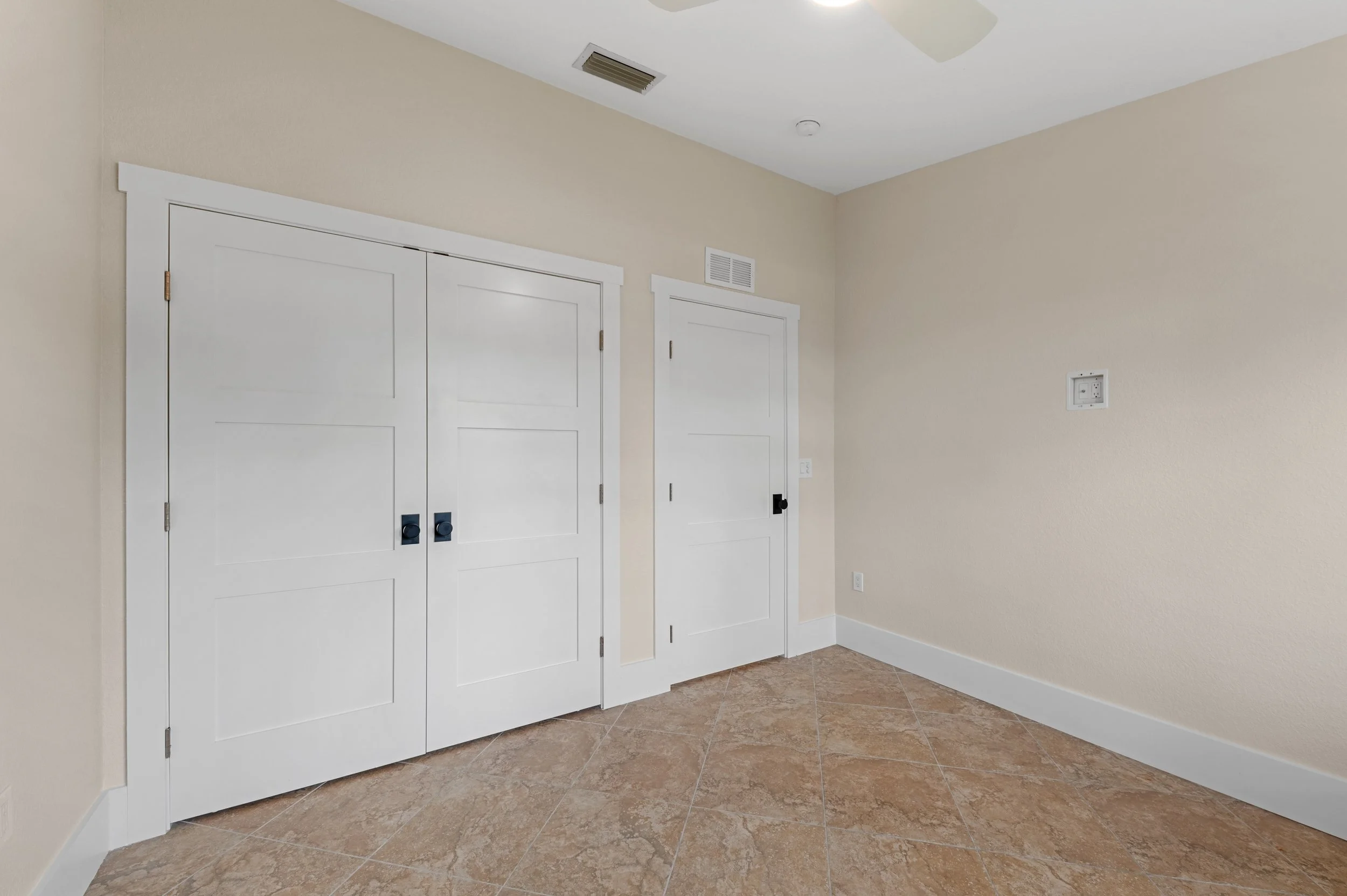 Empty room with beige walls, white trim, beige tiled floor, and two closed white doors with black handles, one of which has a lock. There is a vent near the ceiling and a small wall-mounted electrical panel.
