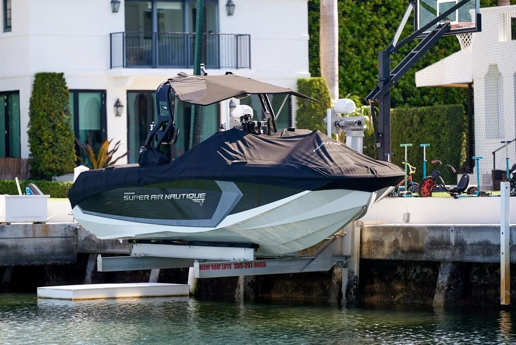 A white and black Super Air Nautique motorboat covered with a black cover, docked at a waterfront with a residential building and several outdoor toys in the background.