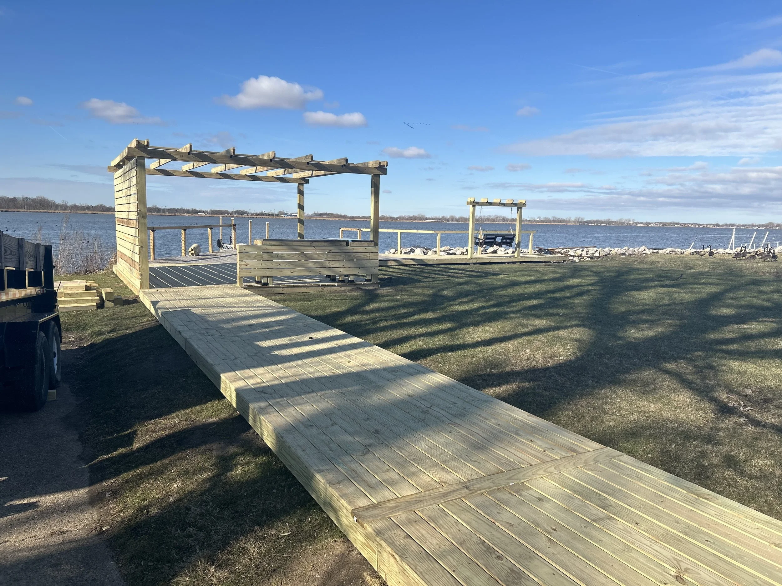 A wooden dock extending over the water with a pergola structure on the left and a second smaller structure further back, near a rocky shoreline under a partly cloudy sky.