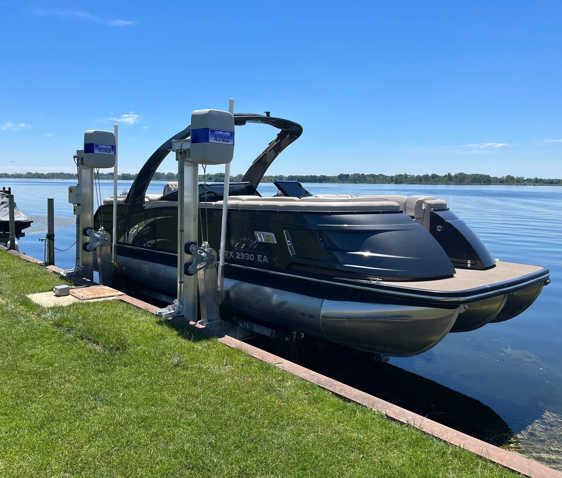 A black motorboat docked at a pier on a calm lake, with a clear blue sky and distant tree line.