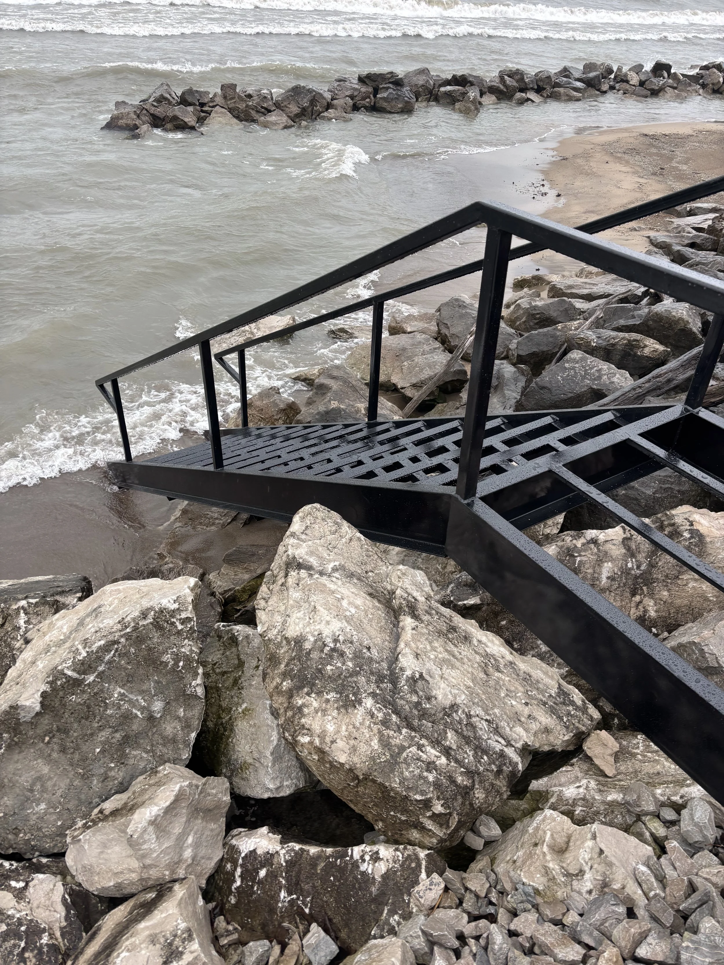 Black metal staircase descending to a rocky shoreline by the water with waves and a rocky breakwater in the background.