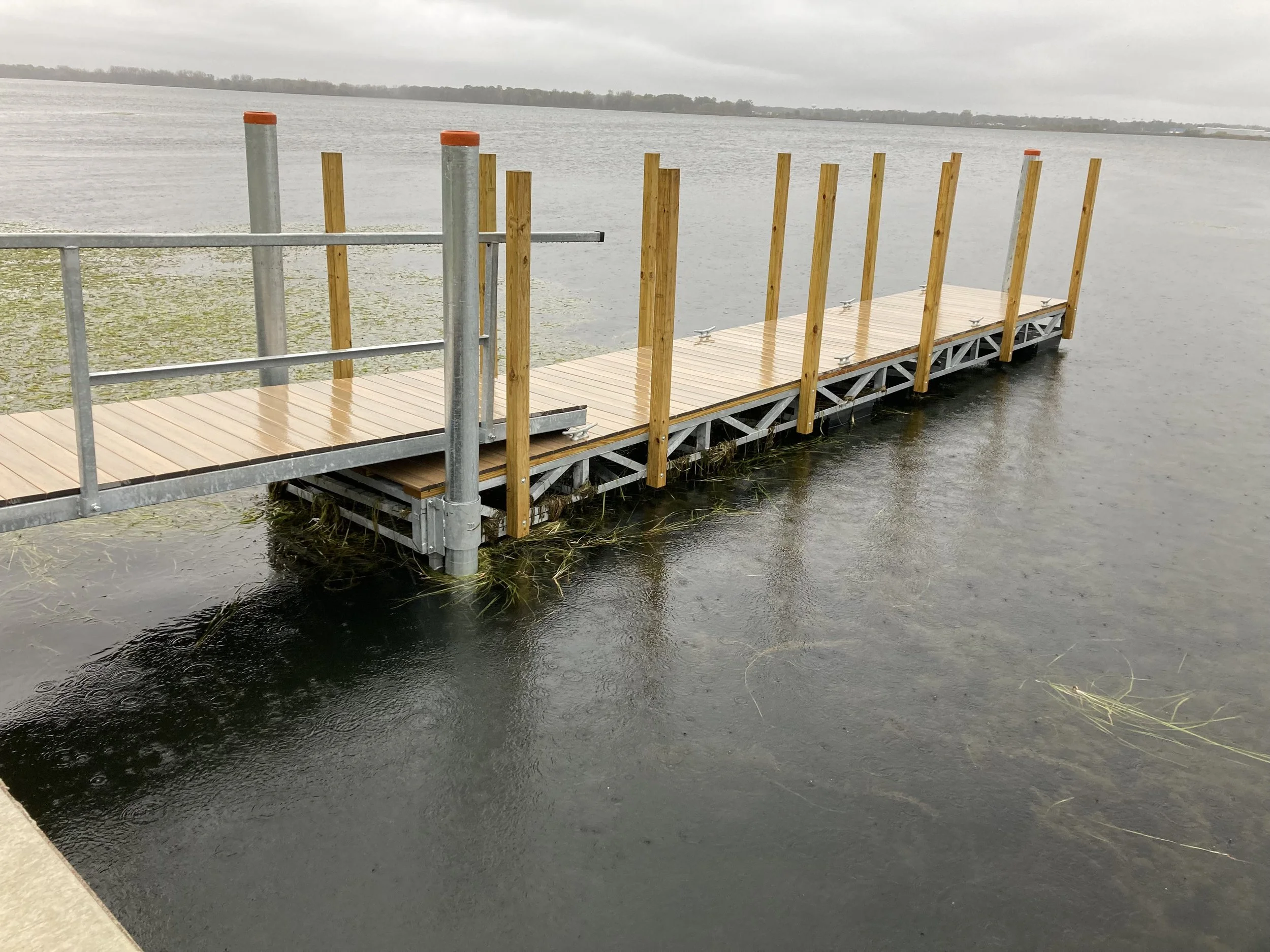 A partially constructed dock with wooden planks and vertical wooden support beams extending into a body of water on a cloudy day.