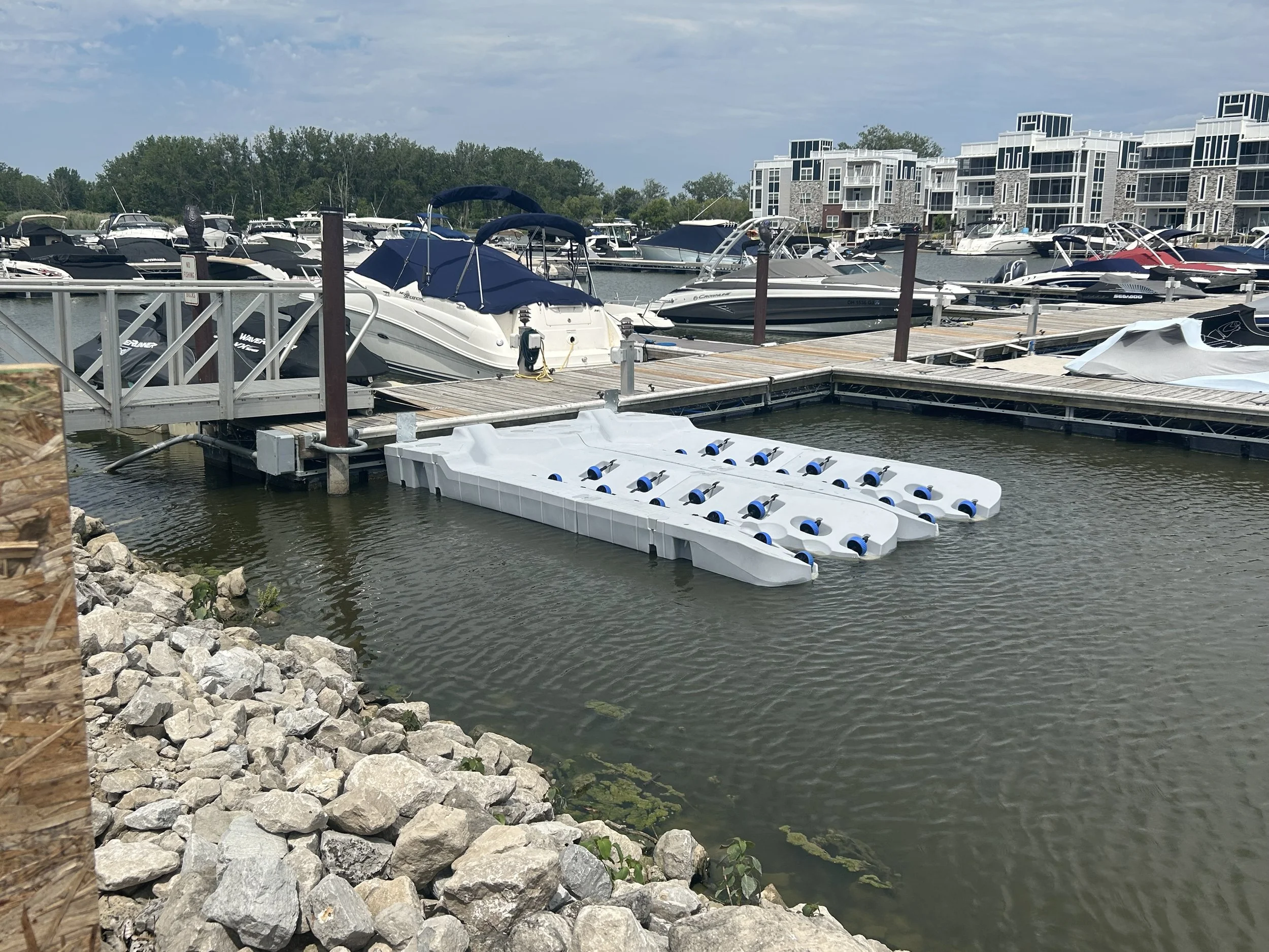 A marina with multiple boats docked, including a white boat with a blue canopy. Floating white paddleboards are attached to a dock. The marina is in a residential area with modern townhouses visible in the background.