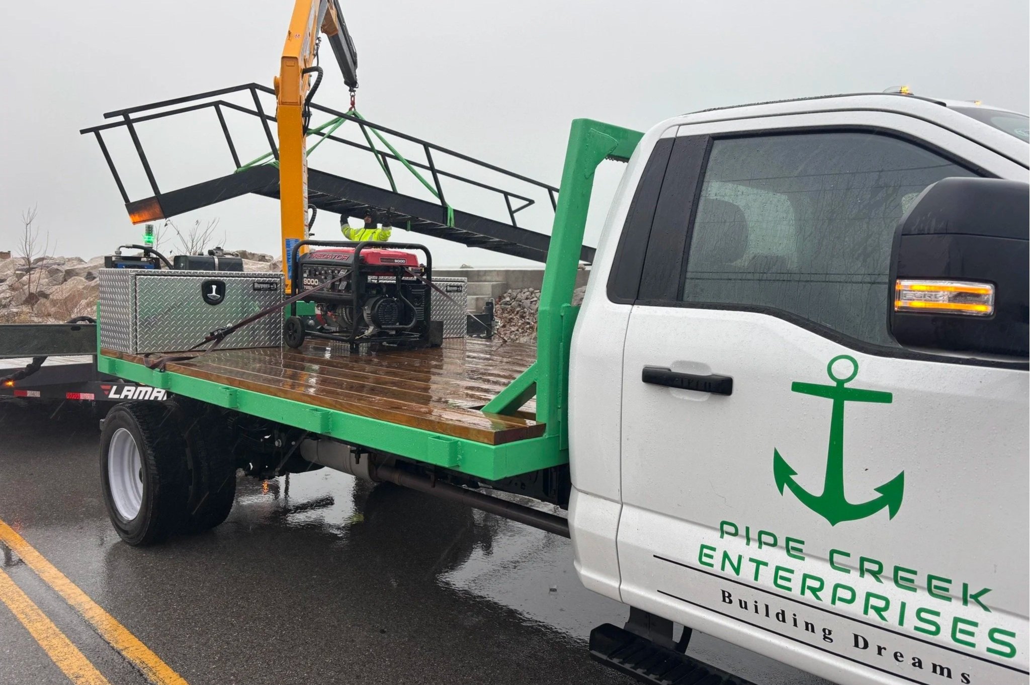 A flatbed truck from Pipe Creek Enterprises transporting a metal staircase or platform, with a construction worker in a yellow jacket and helmet working near the staircase on a rainy day.