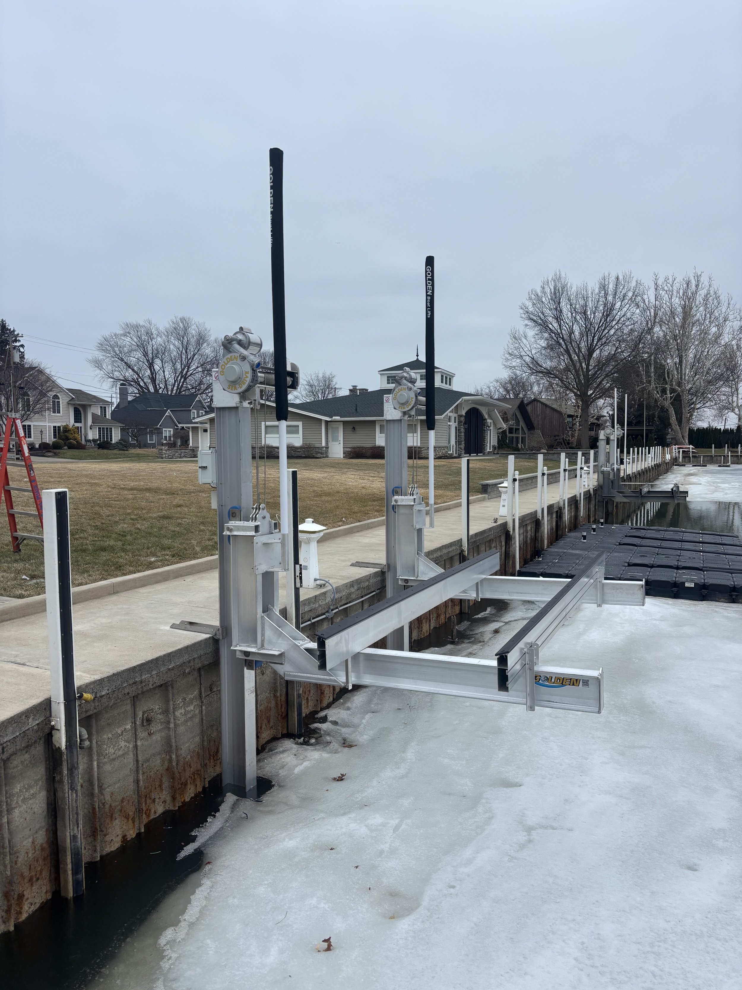A white and black Super Air Nautique motorboat covered with a black cover, docked at a waterfront with a residential building and several outdoor toys in the background.