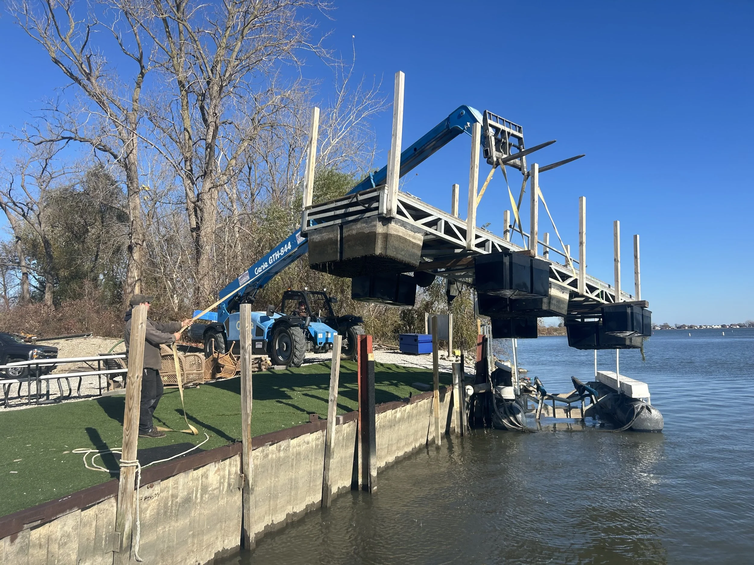 A person is operating a blue crane to lift a wooden platform with railings at the edge of a body of water. The platform is partially submerged with a boat attached beneath it. Tall, leafless trees are in the background under a bright blue sky.