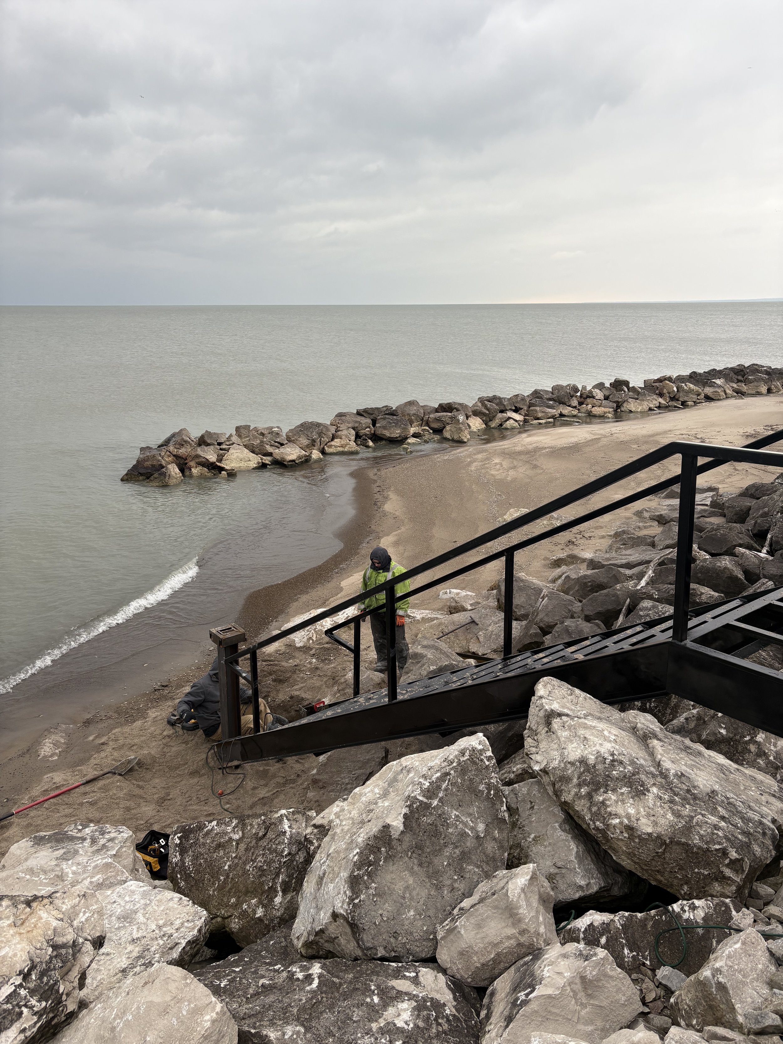 Two workers installing or repairing a metal staircase leading down to a beach with sand and rocks, near a breakwater made of large rocks, under a cloudy sky.