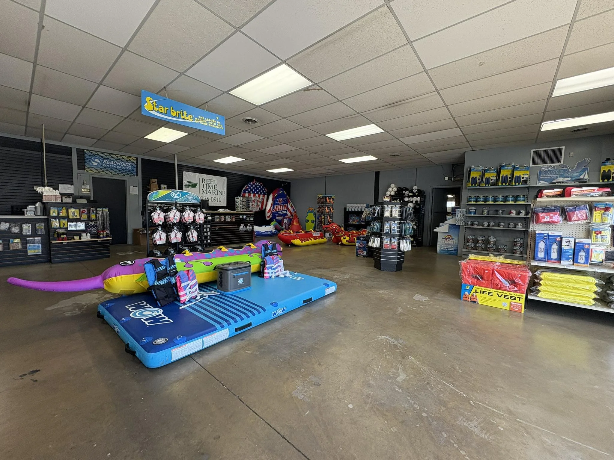 Empty indoor store with various beach and water sports equipment on display, including an inflatable float, life vests, and accessories, with shelves of marine supplies and merchandise.