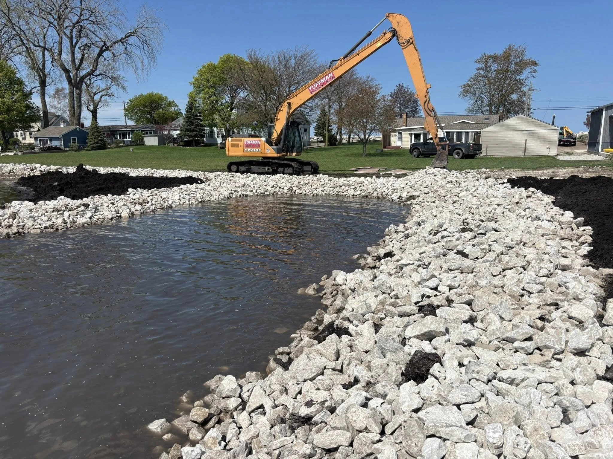 Construction site with a yellow excavator working by a small body of water, surrounded by rocks and soil, in a residential area with houses, trees, and a clear blue sky in the background.