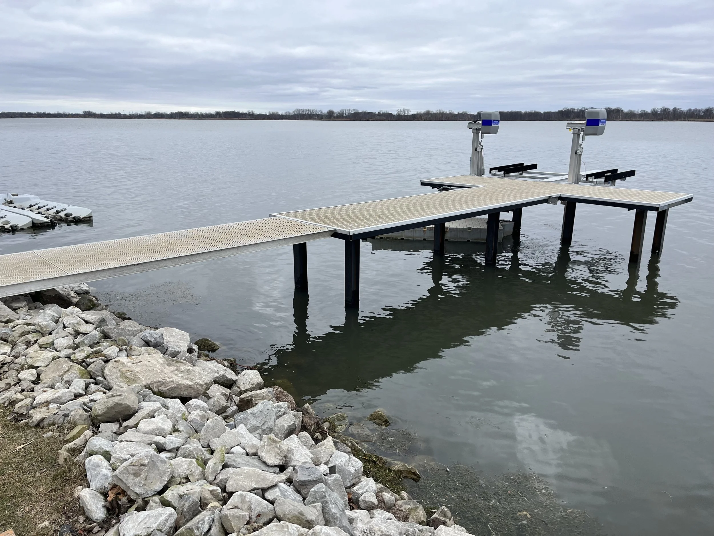 A boat dock with a floating platform extending into a body of water, featuring two boat lifts on the right side and rocks along the shoreline; the sky is overcast.
