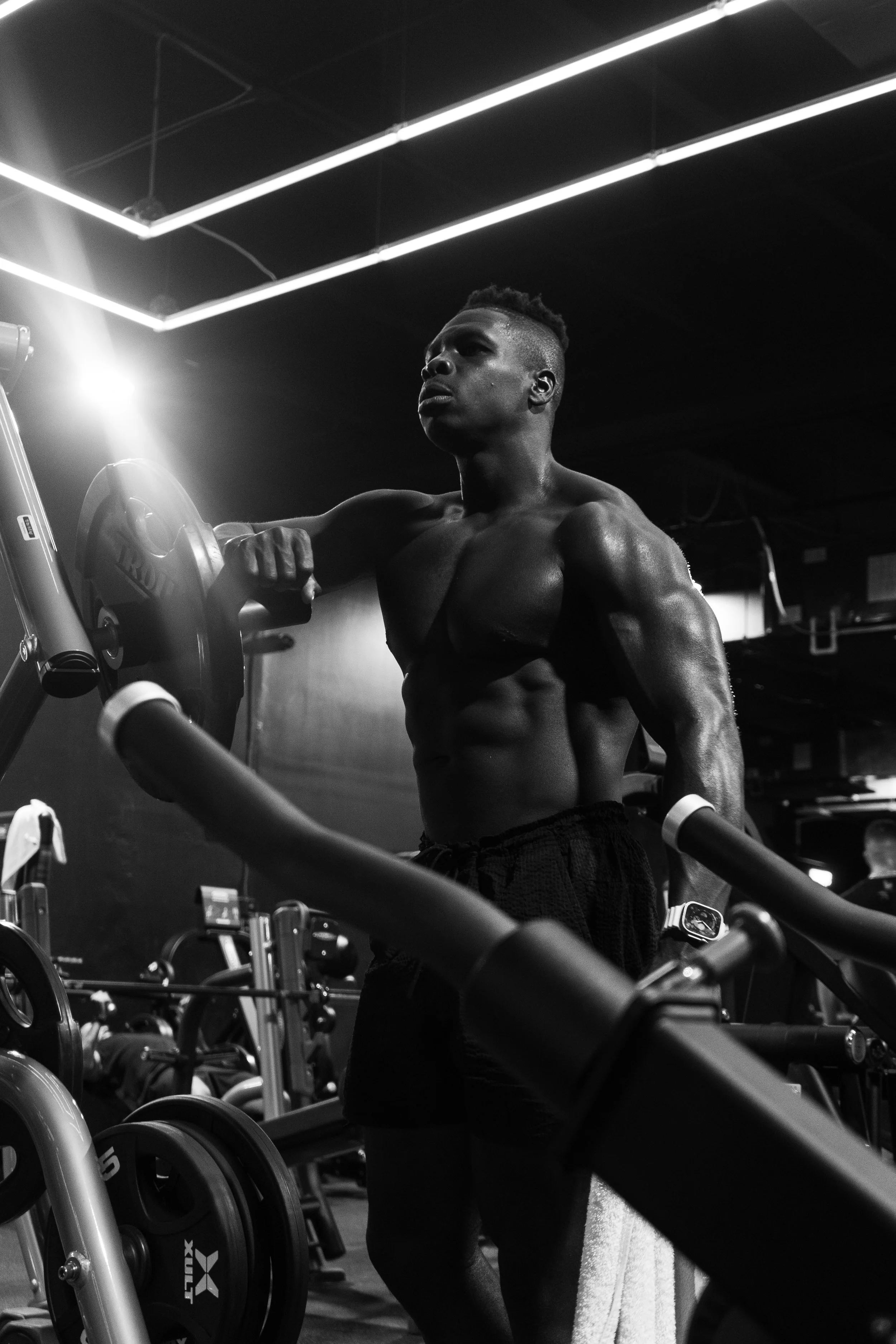 A shirtless man in athletic shorts lifting a dumbbell in a gym with black walls, fluorescent lights, and various exercise equipment.