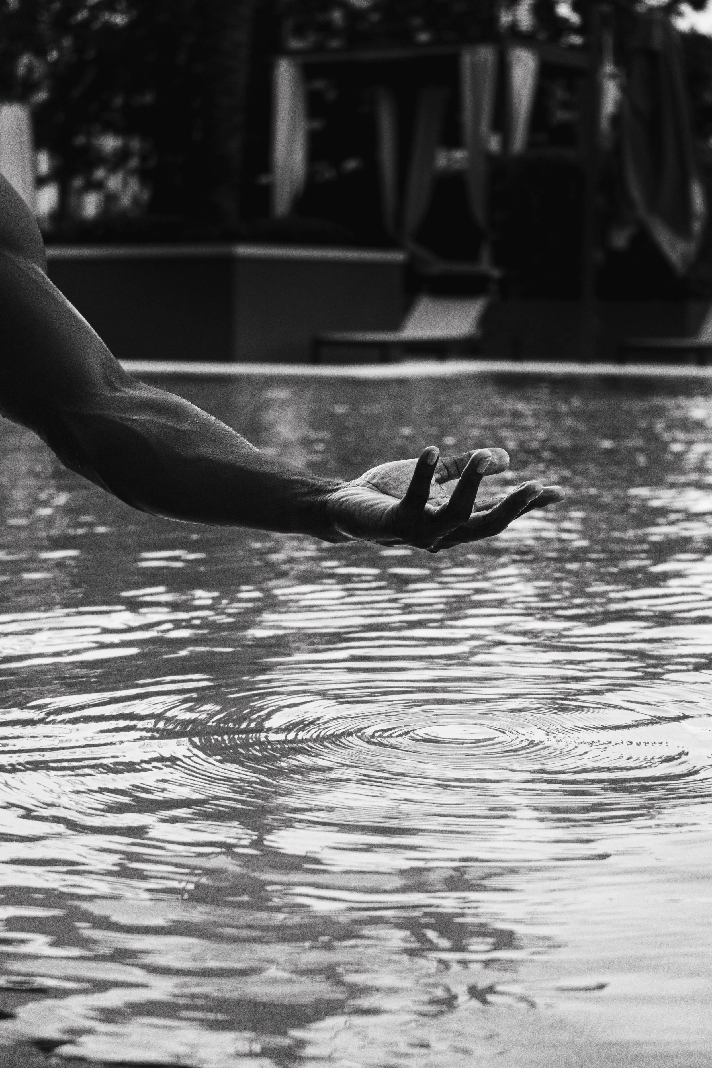 Close-up of a hand with extended fingers above rippling water, with a blurred background showing trees and a structure.
