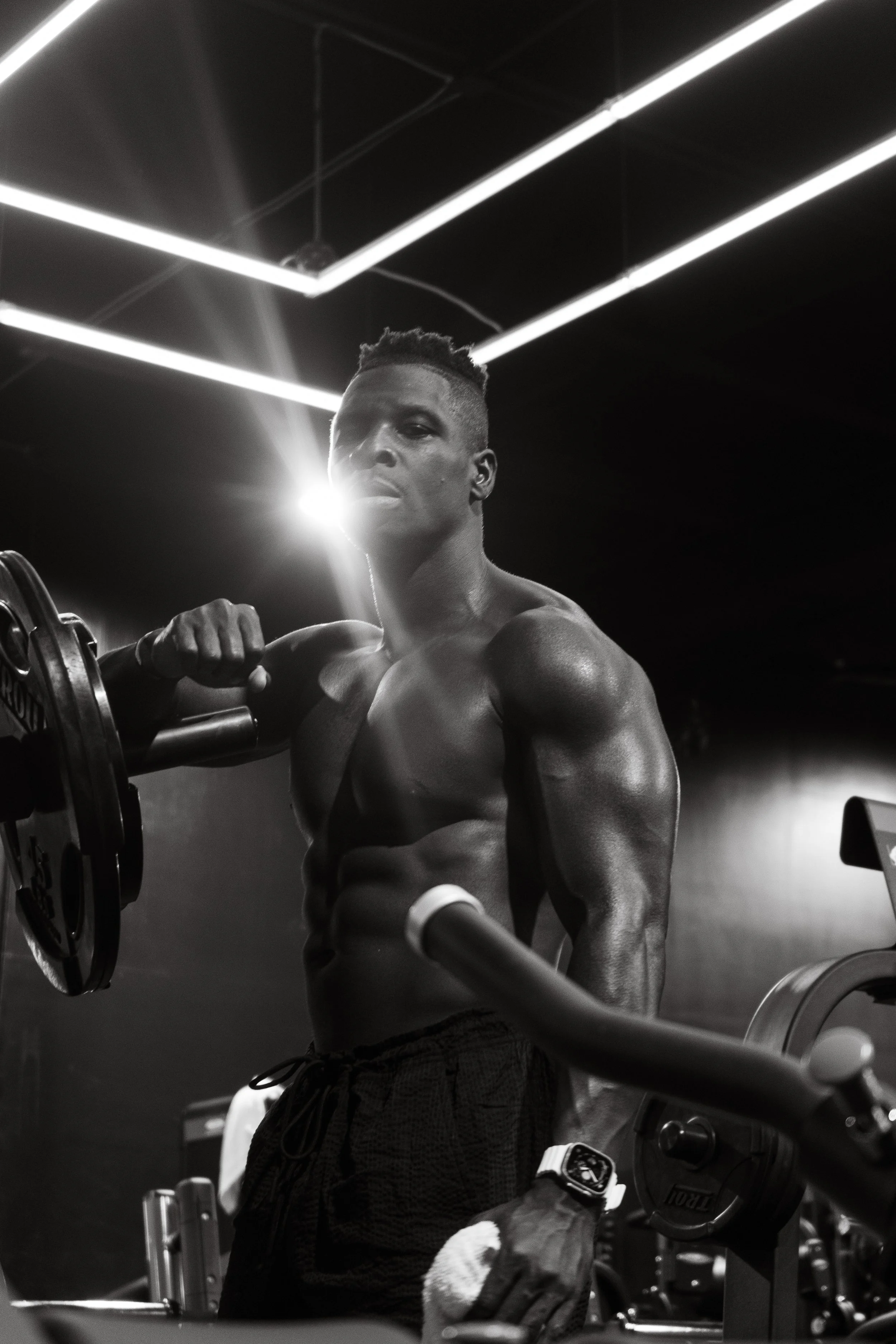 A muscular man with a tattoo on his right arm, wearing a watch and gloves, working out in a gym with LED lights on the ceiling.