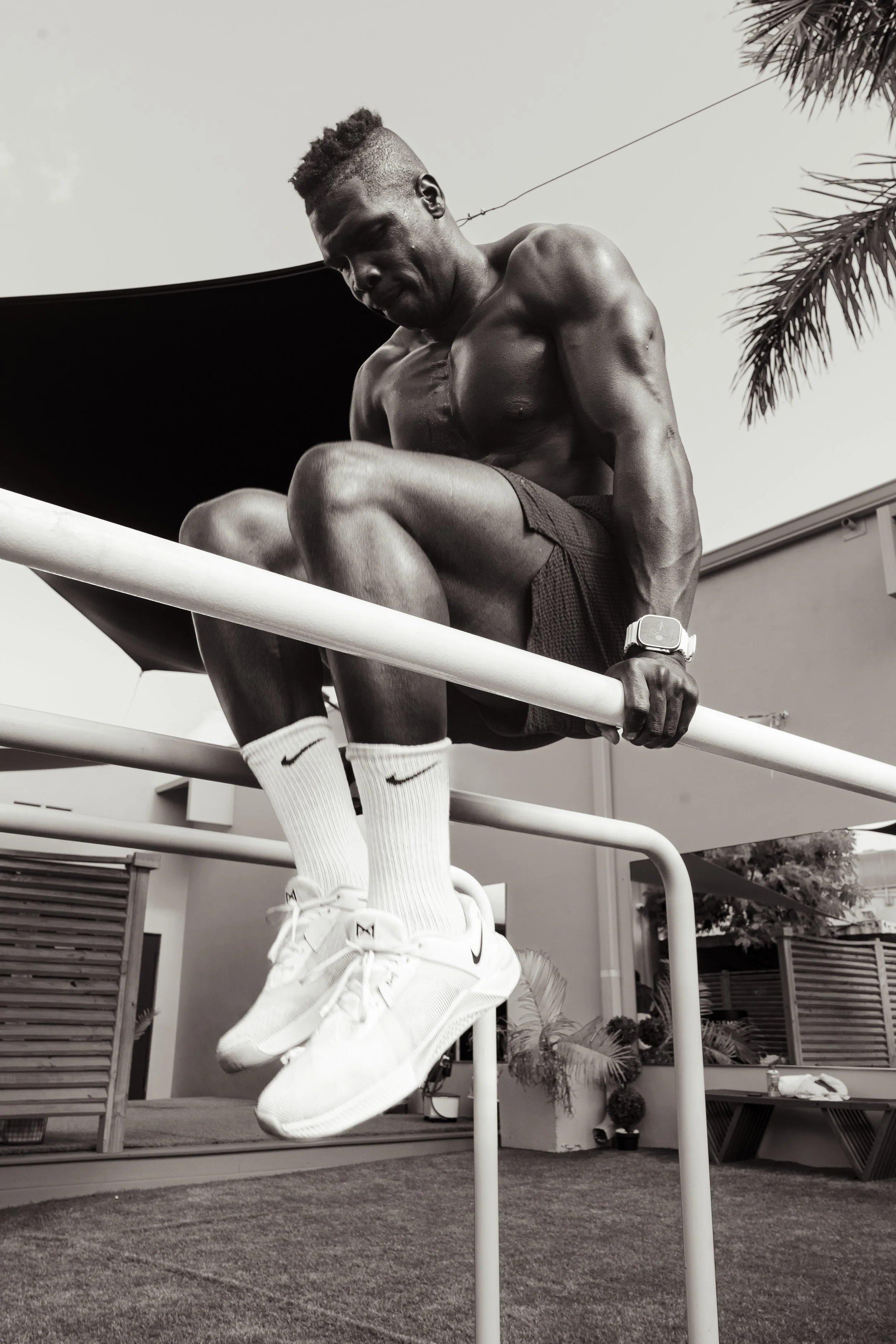 A shirtless young man with a muscular build is sitting on a playground apparatus, with his legs bent and holding onto the bars. He is wearing a watch, Nike socks, and sneakers, with trees in the background and buildings nearby.