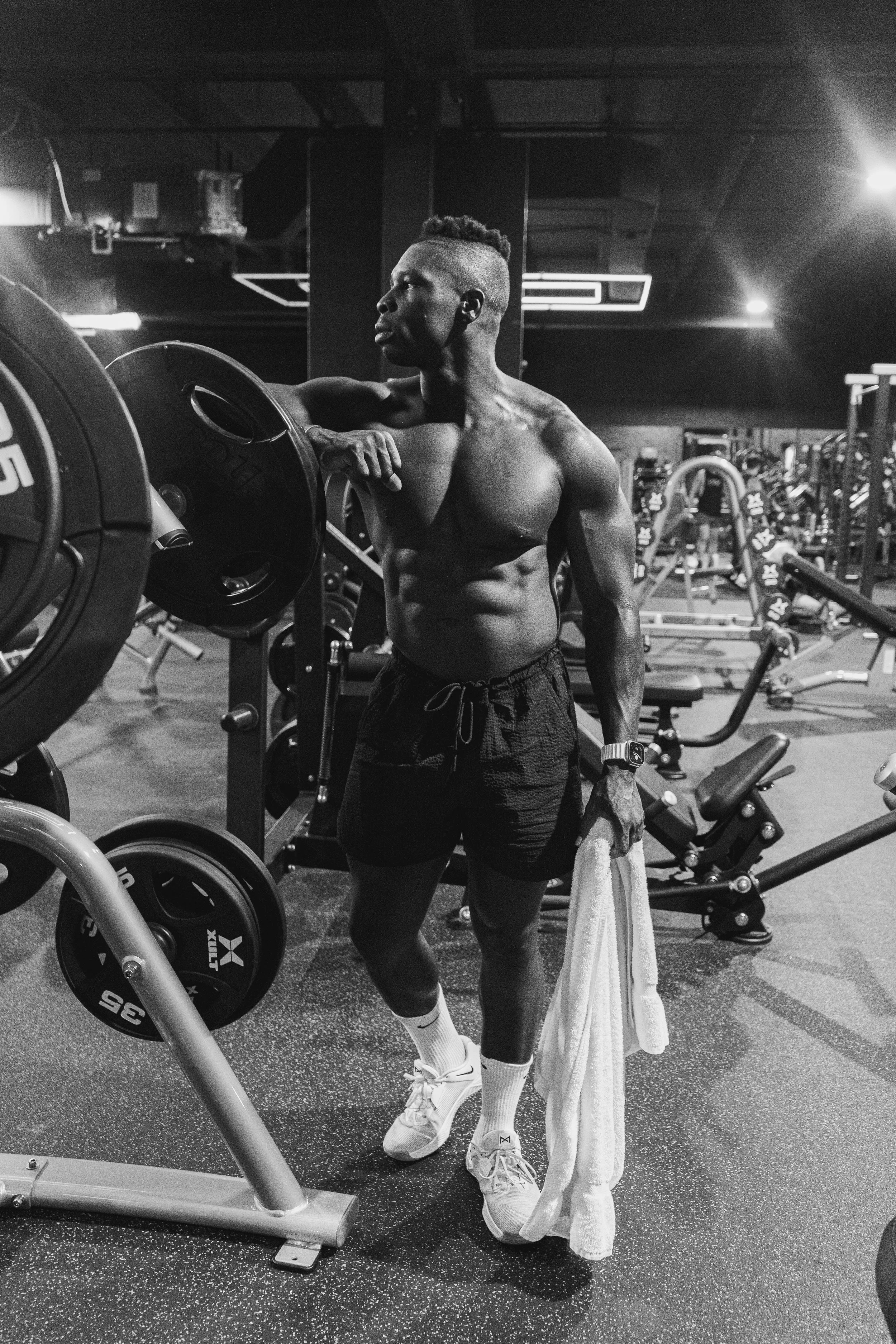 A shirtless man with a mohawk hairstyle, wearing shorts, white socks, and sneakers, standing in a gym. He is holding a towel and leaning on a piece of gym equipment, looking to the side. The gym has various machines and weights in the background, with bright overhead lights and a dark ceiling.