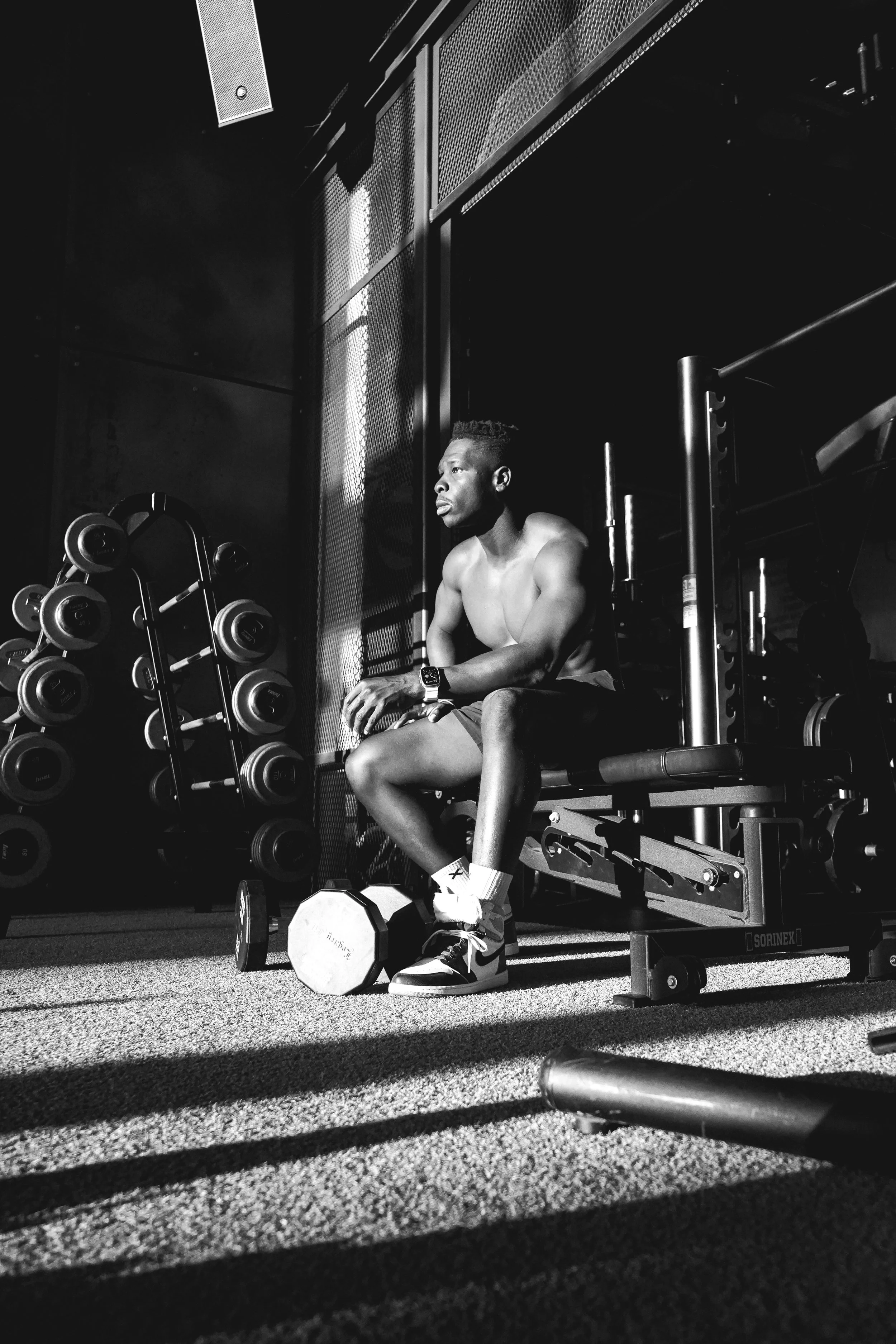 A shirtless man sitting on a workout bench at the gym, resting with a dumbbell on the floor beside him, surrounded by exercise equipment and weights.