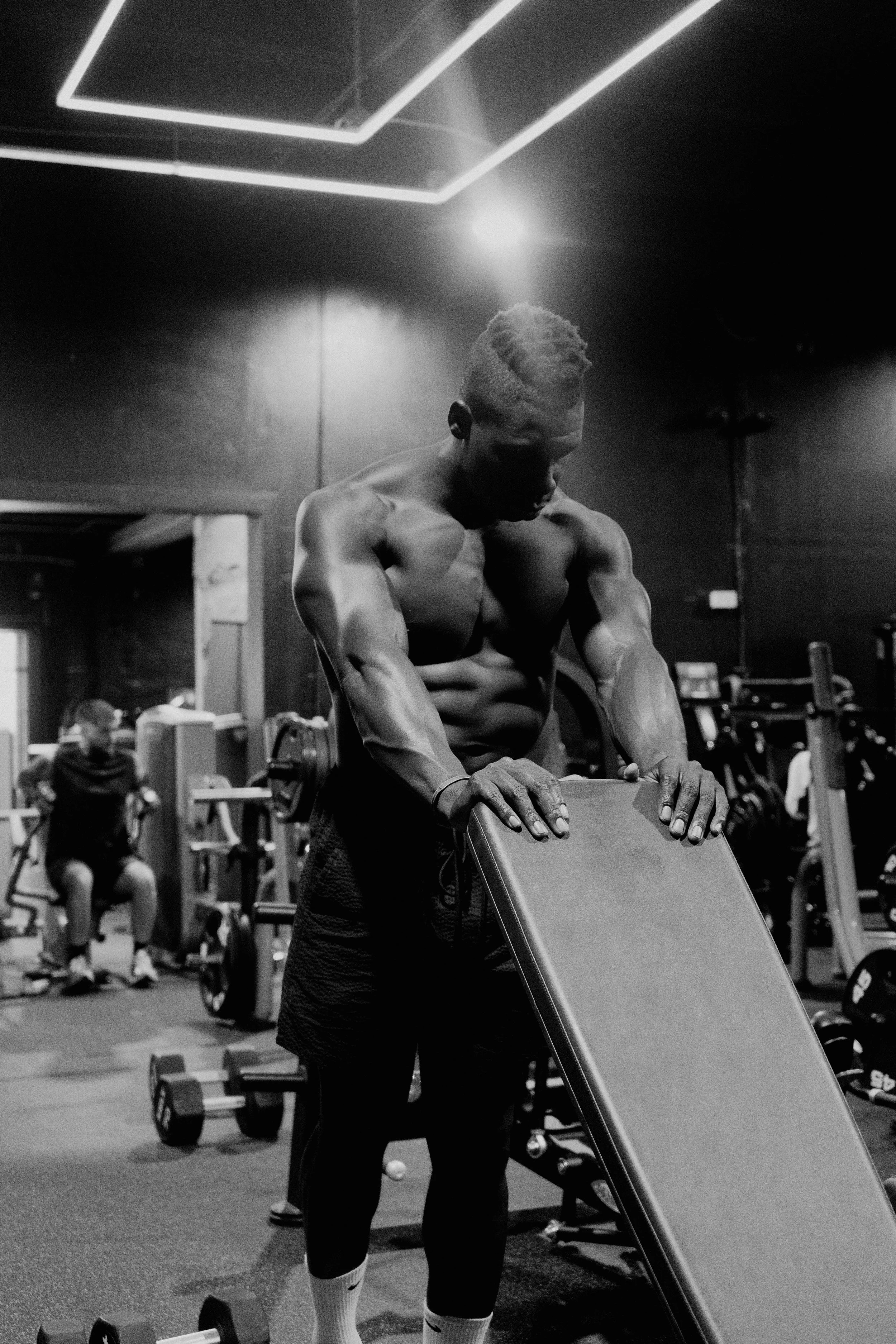 A muscular man with a short hairstyle is doing a plank exercise on a raised surface in a gym. We can see other gym equipment and people working out in the background.