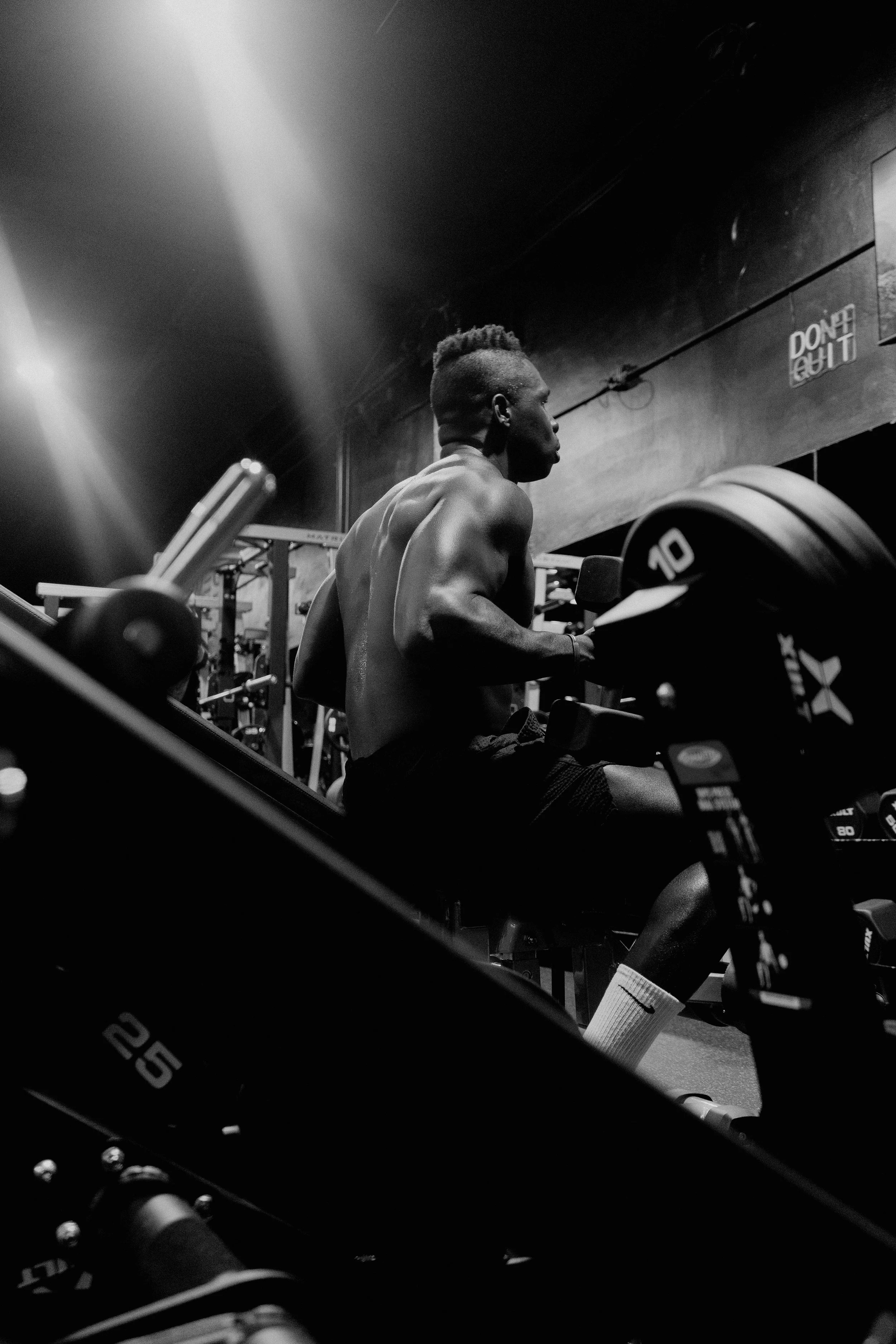 A muscular man with a mohawk hairstyle working out in a gym, sitting on a bench, and surrounded by weightlifting equipment in a black and white photo.