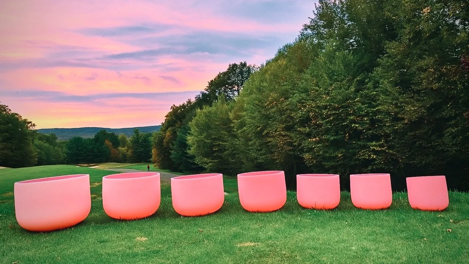 Six pink illuminated singing bowls on a green field during sunset, with trees and distant hills in the background.