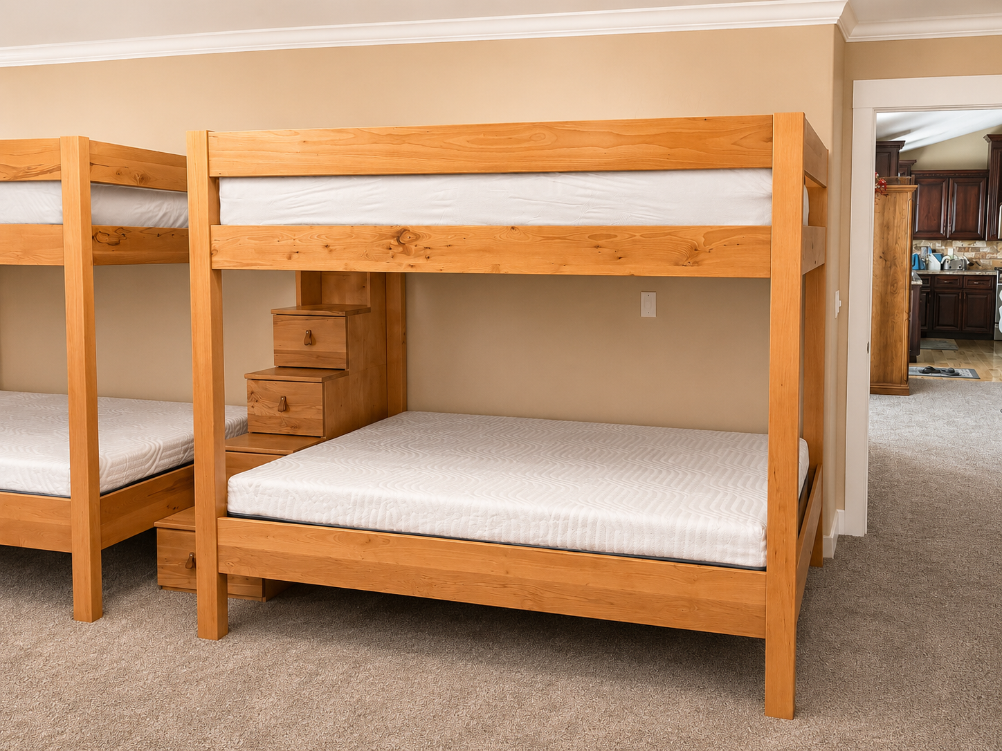 Wooden bunk beds with mattresses, next to a small bedside table with drawers, in a beige-walled room with carpeted floor, and a view into a kitchen with dark cabinets in the background.