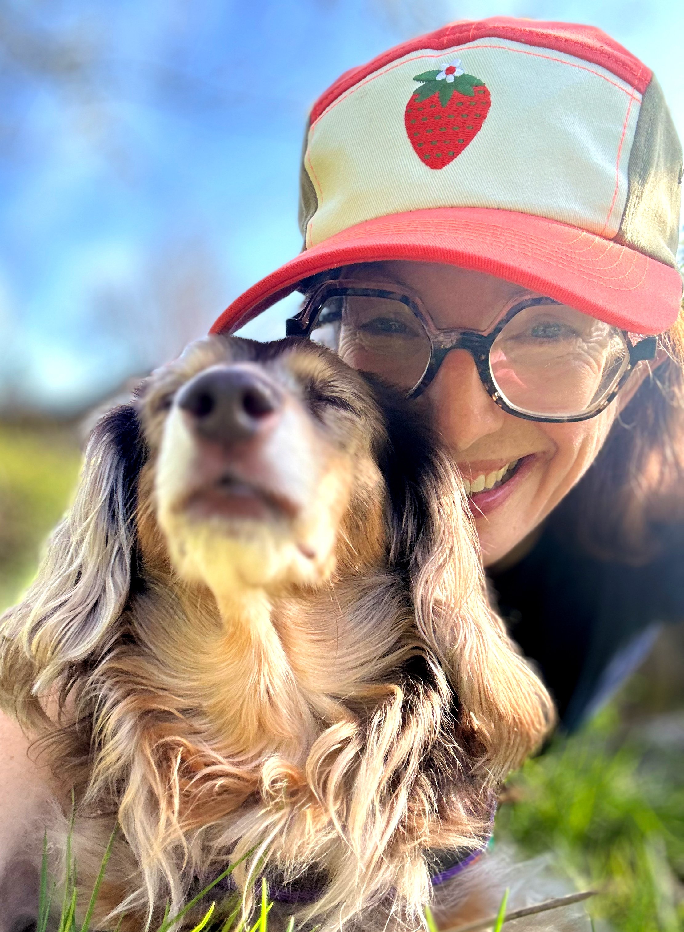 A smiling woman in glasses and a strawberry hat with a dachshund dog outdoors.