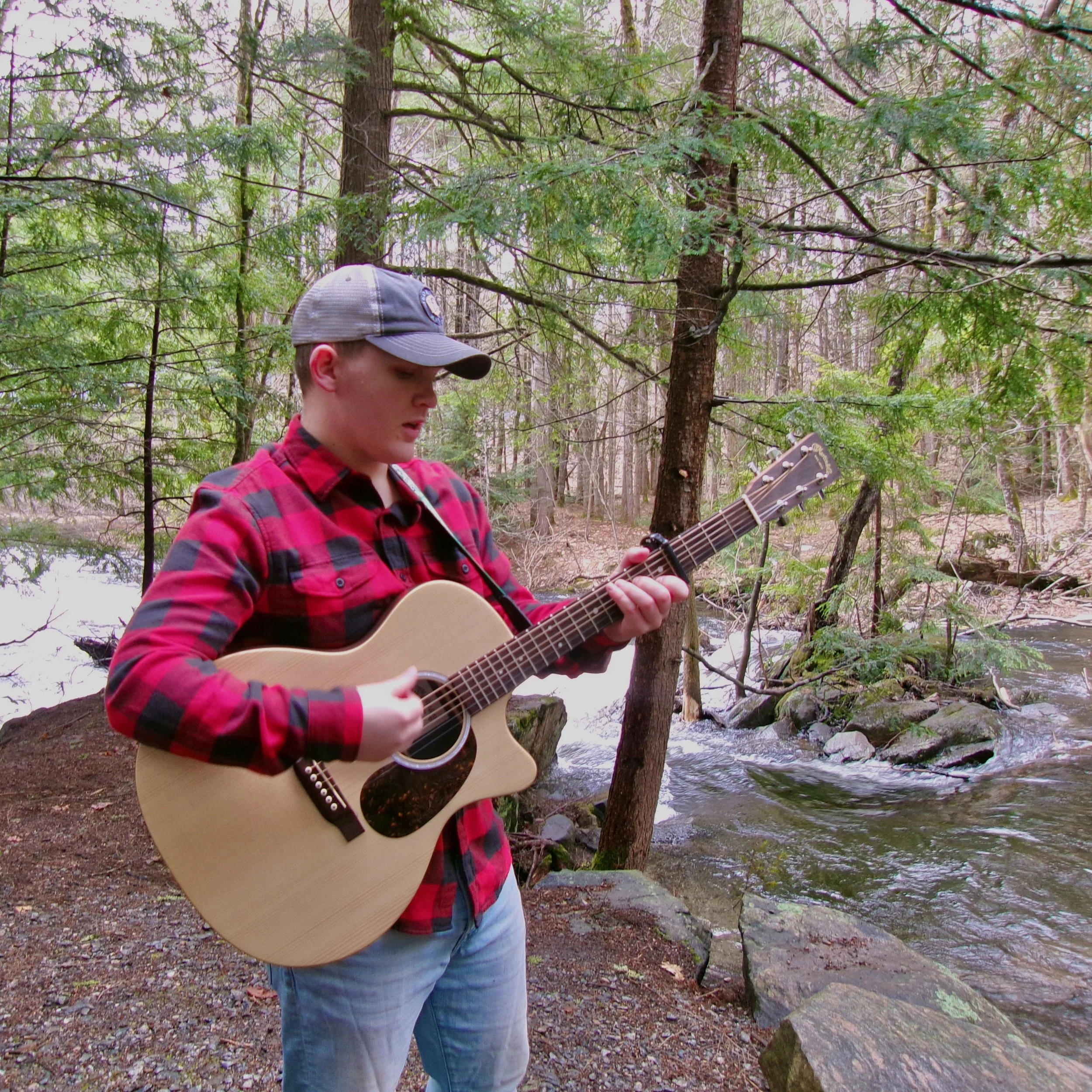 A young man wearing a gray cap and red plaid shirt plays an acoustic guitar outdoors near a flowing stream surrounded by trees in a forest setting.