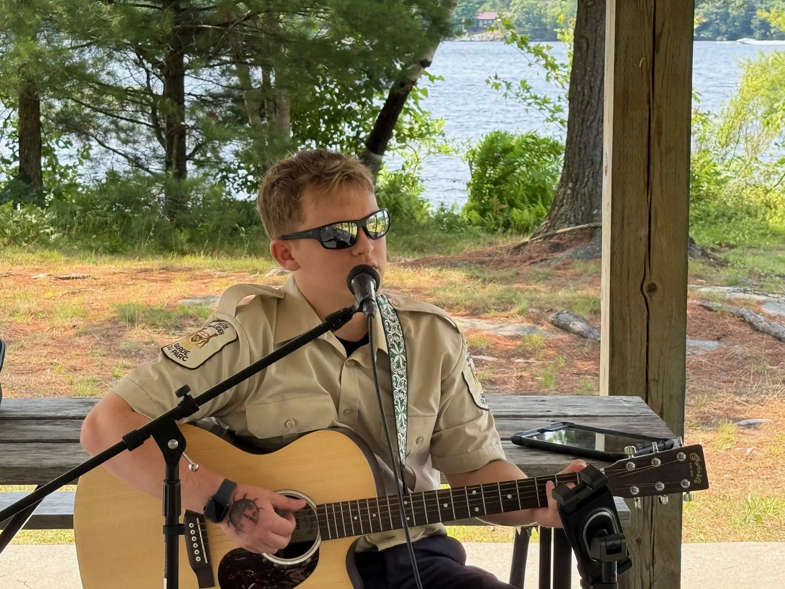 A person in a tan shirt with badges, sunglasses, and tattoos plays an acoustic guitar and sings into a microphone at an outdoor event near a lake, with trees and water in the background.